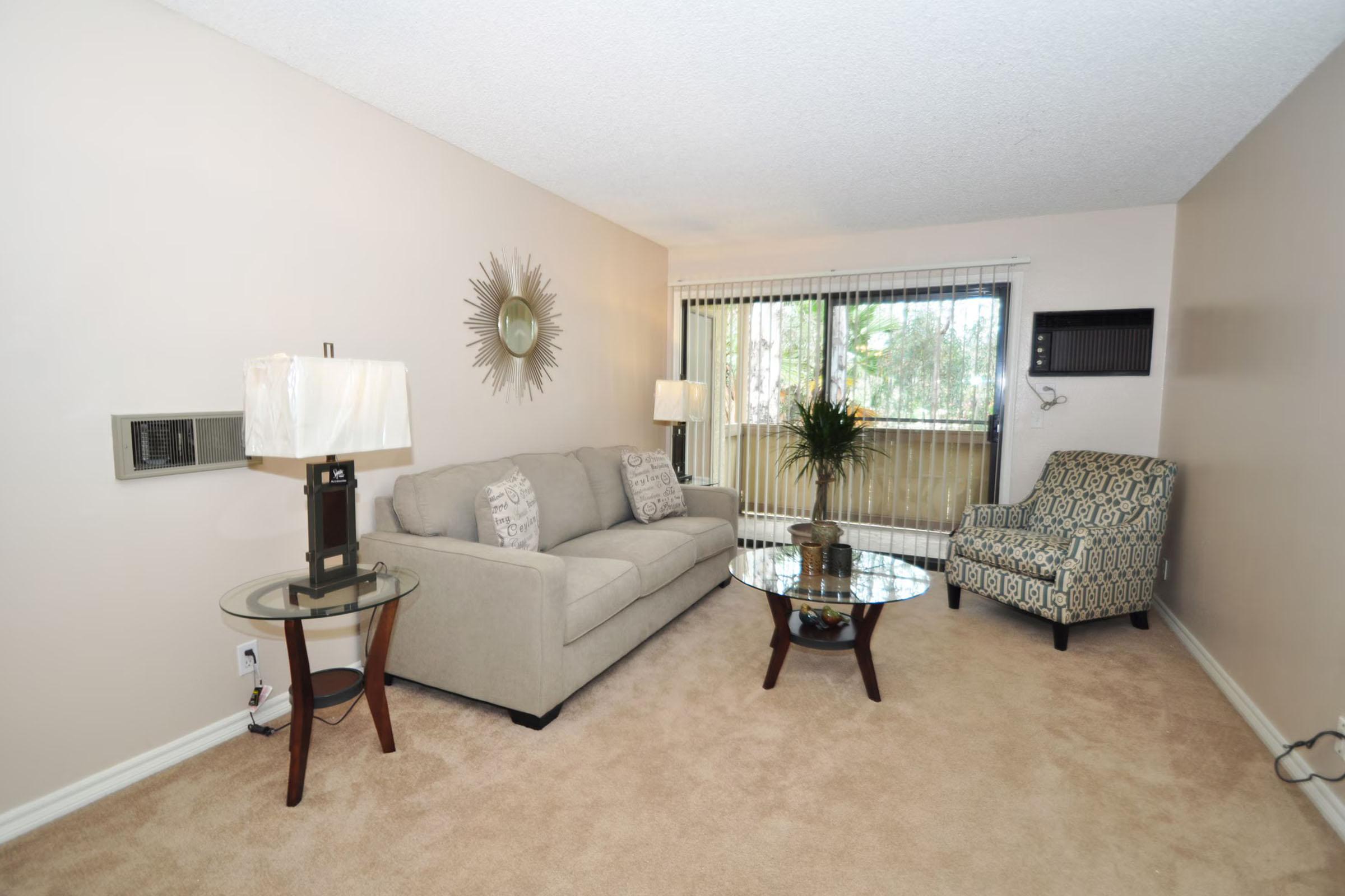 A cozy living room featuring a light beige sofa with decorative pillows, a glass coffee table, and a patterned accent chair. There’s a table lamp on a small wooden side table, and a large mirror on the wall. Natural light enters through a window, complemented by indoor plants. The carpet is light-colored, adding warmth to the space.