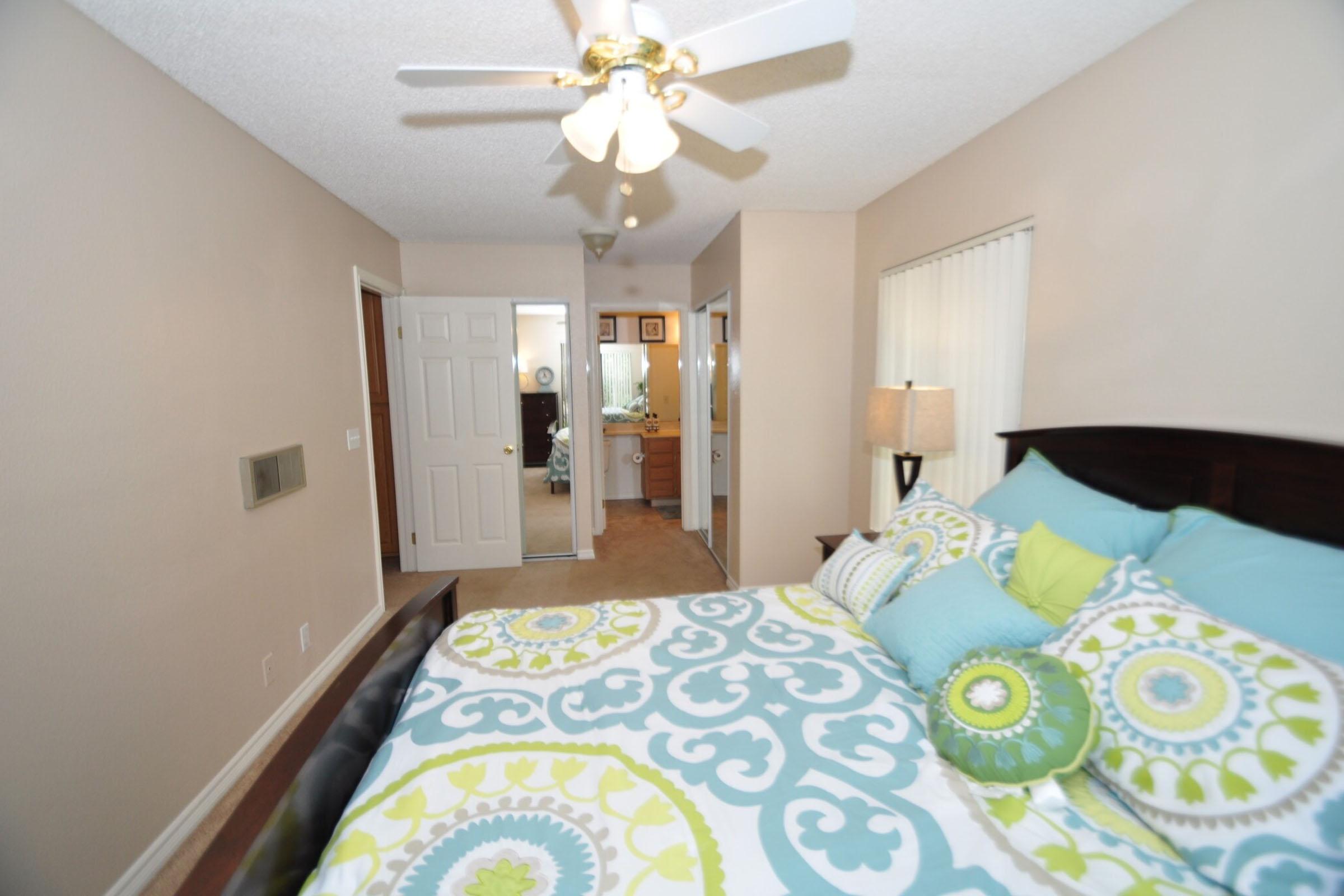 A brightly lit bedroom featuring a bed with colorful bedding in turquoise and lime green. There is a decorative pillow on the bed, a ceiling fan, and light-colored walls. A door leads to a bathroom area visible in the background. The flooring is carpeted, and natural light comes in through a window.