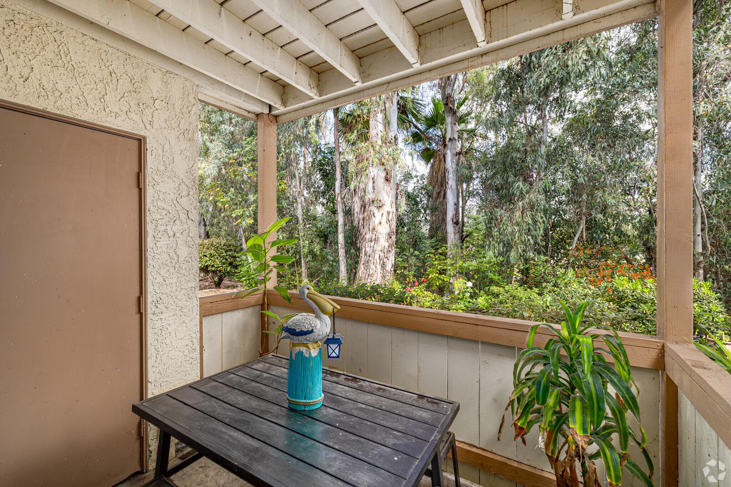A small outdoor balcony with a wooden table and a decorative blue lantern, surrounded by green plants. In the background, tall trees provide a natural, serene view. The balcony has a beige wall and a door to the side, creating a cozy, inviting atmosphere.