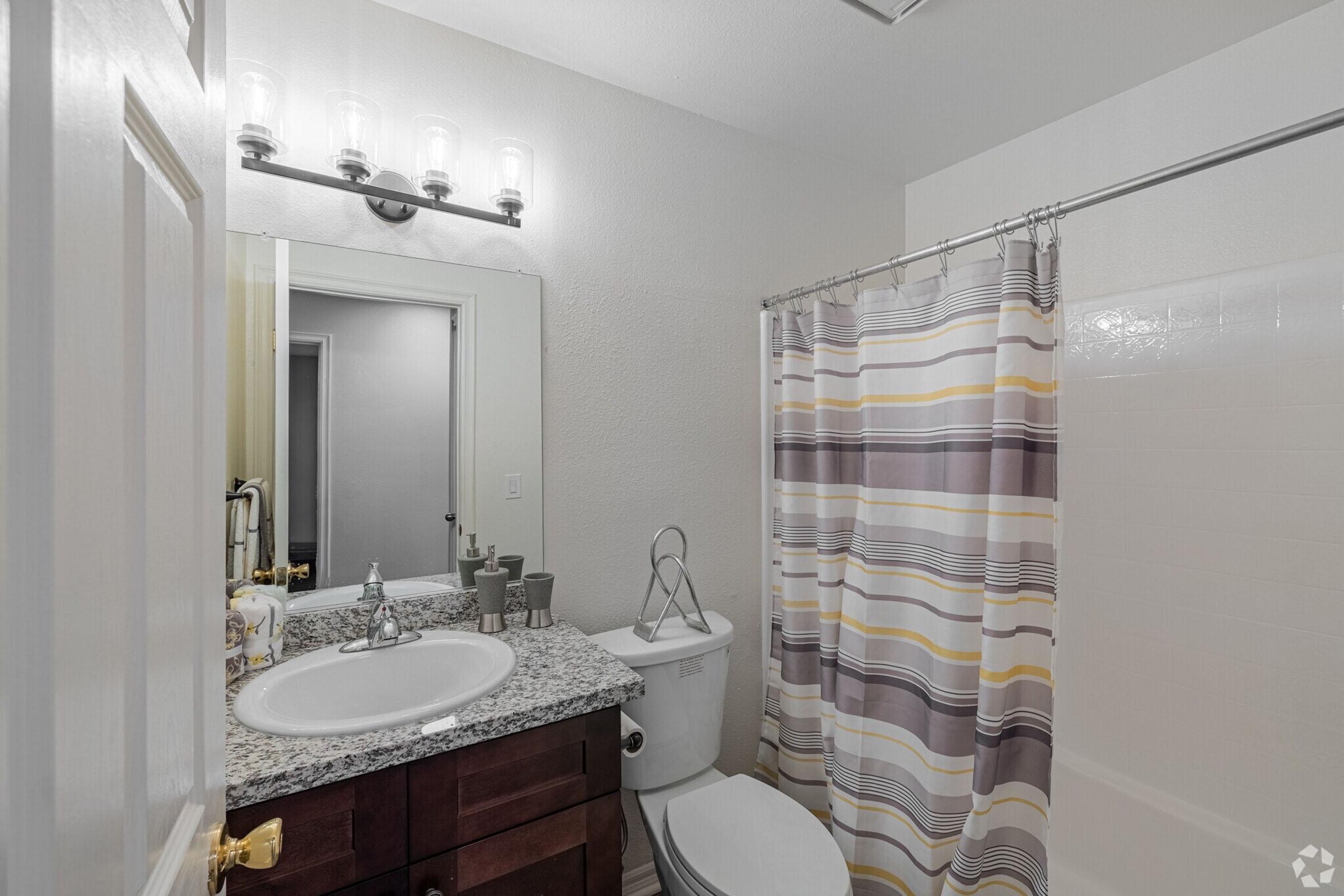A well-lit bathroom featuring a sink with a granite countertop, a mirror, and a shower with a striped curtain. Accessories like toiletries are neatly arranged on the counter, and a bath towel rack is visible. The overall decor is modern and clean.