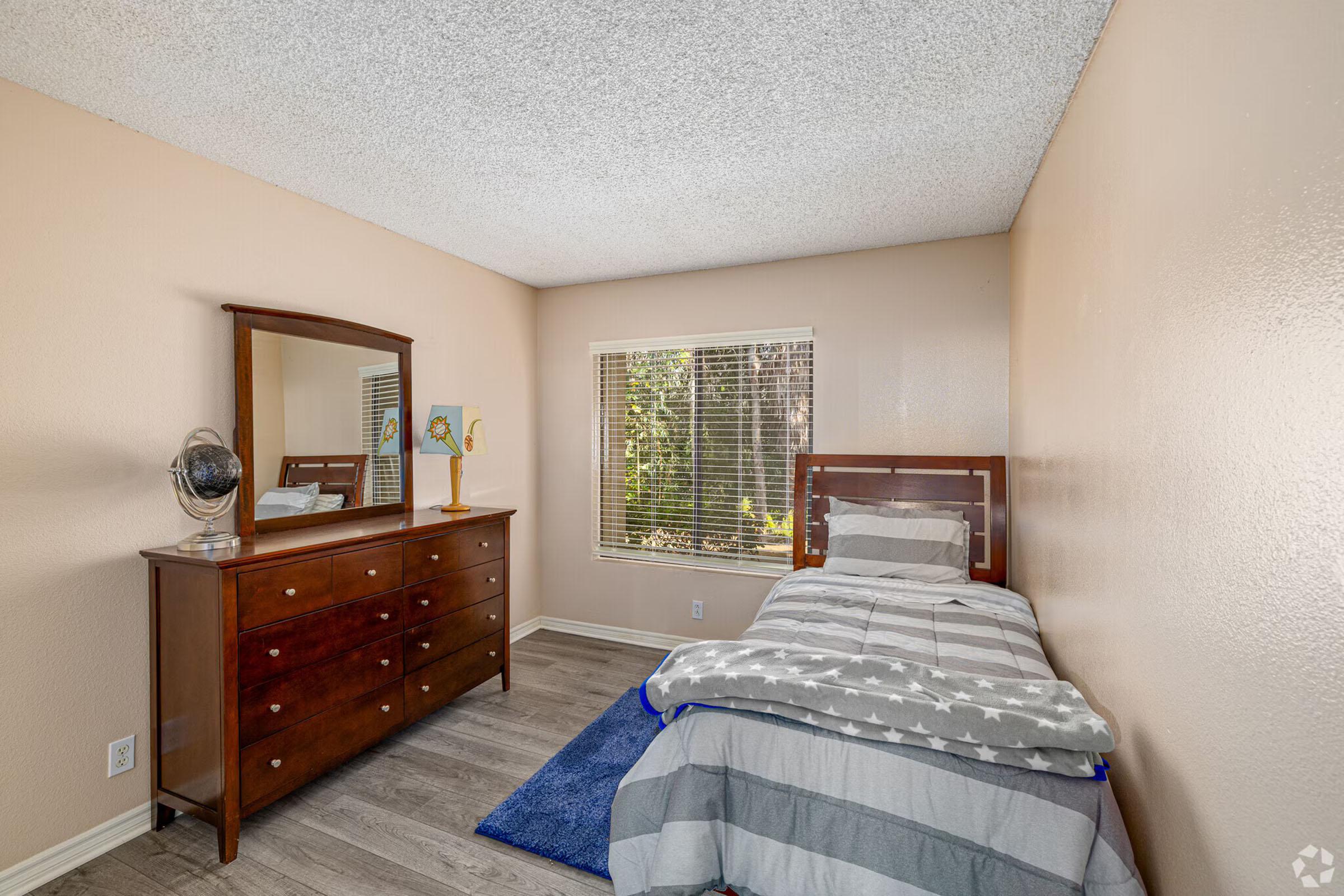 Cozy bedroom featuring a single bed with gray and white bedding, a wooden dresser with a large mirror, and a blue area rug. Natural light streams in from a window with blinds, adding warmth to the neutral-colored walls. A globe decorates the dresser, enhancing the room's character.