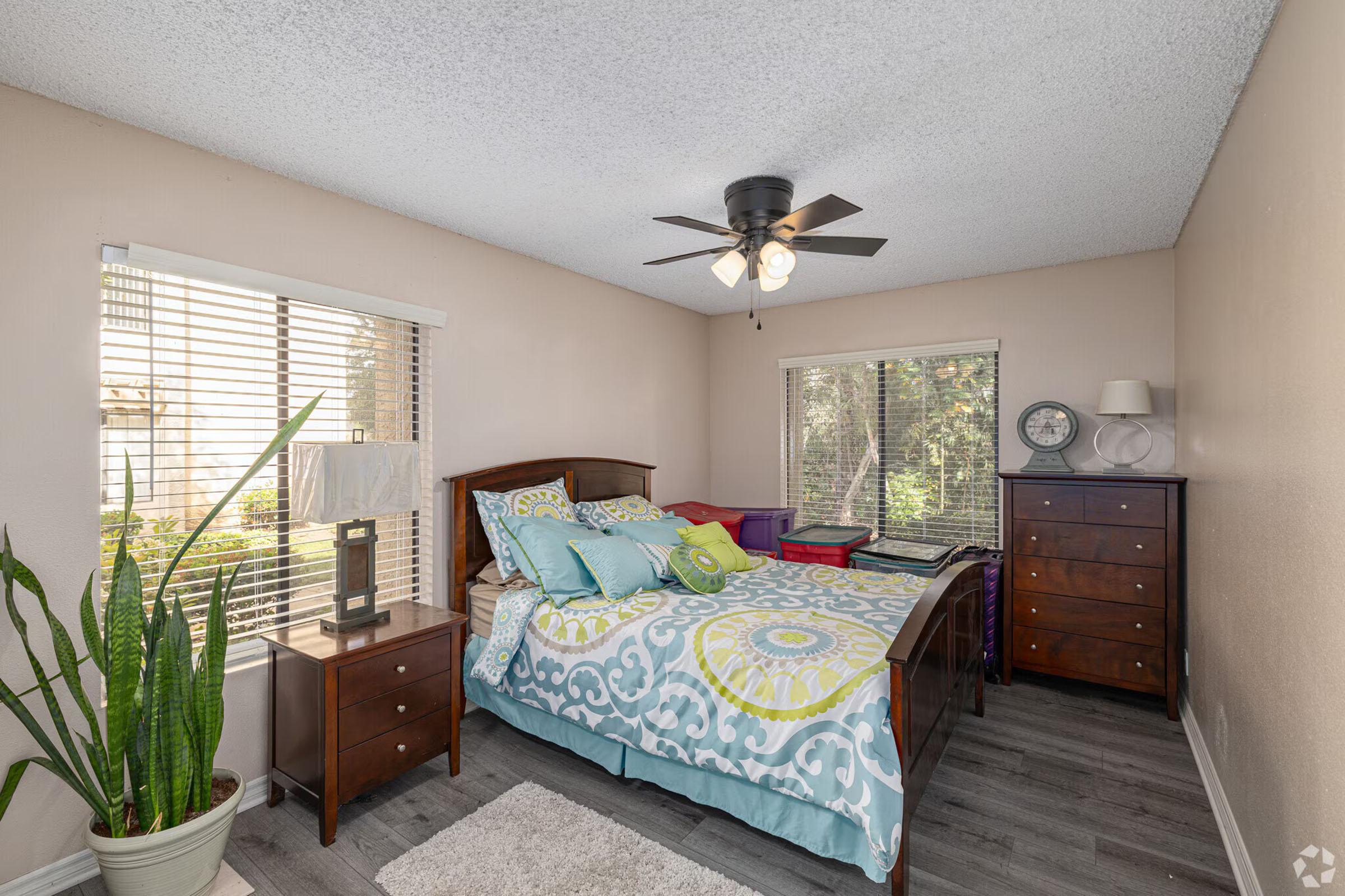 A cozy bedroom featuring a wooden bed with colorful bedding, a bedside lamp, a dresser, and a large window with blinds. A potted plant adds a touch of greenery, and there’s a small table with decor next to the window. The walls are painted in a soft color, creating a warm and inviting atmosphere.