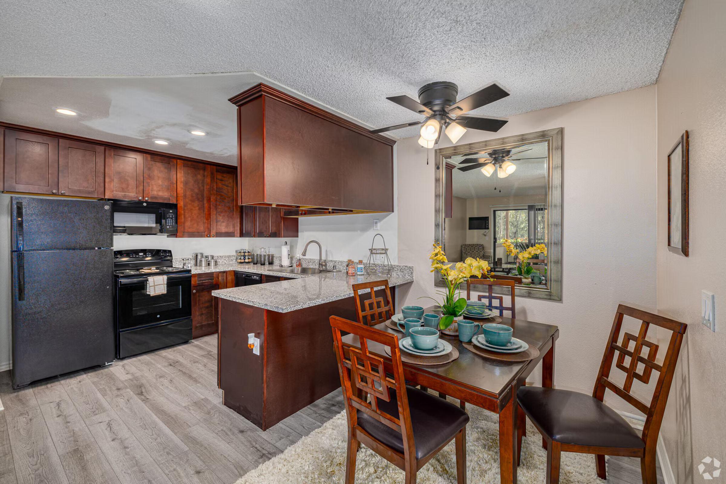 A cozy kitchen and dining area featuring dark wood cabinets, granite countertops, and a black refrigerator and stove. A round dining table is set with plates and cups, surrounded by four wooden chairs. Bright yellow flowers adorn the table, and a ceiling fan adds to the space's warmth.