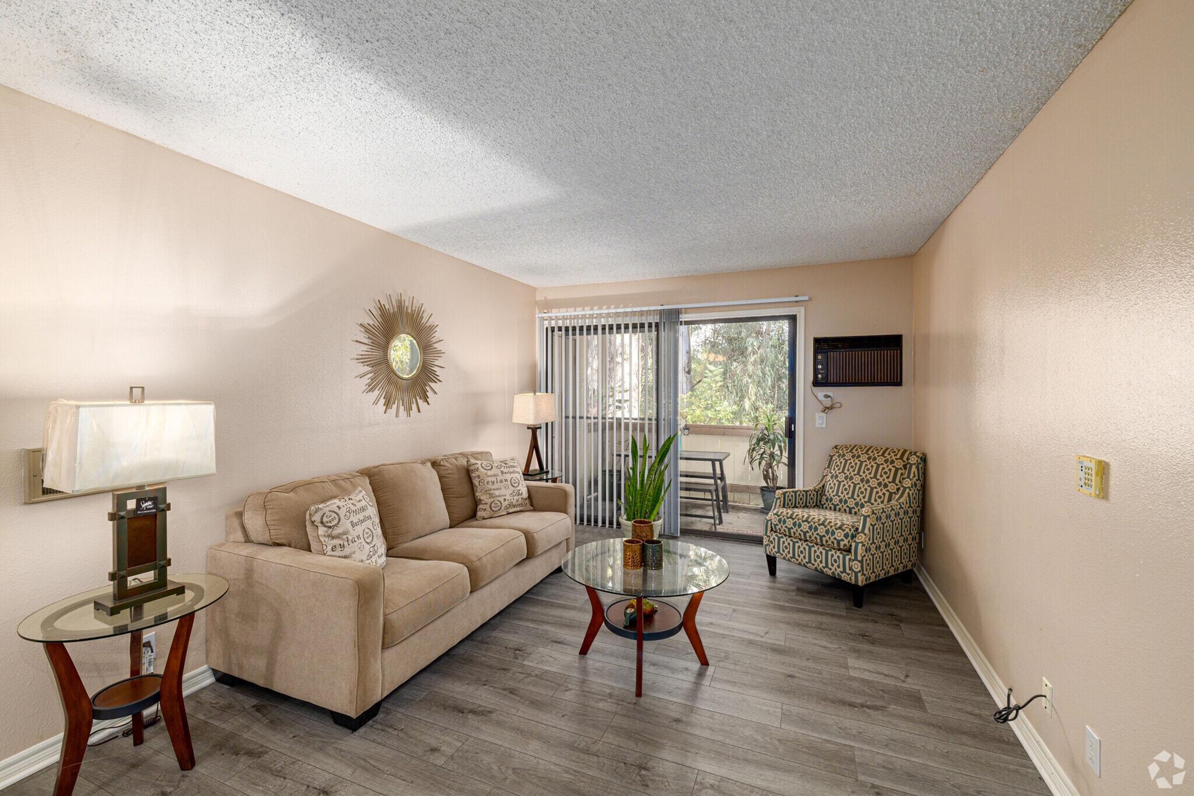 A cozy living room with a beige sofa and a glass coffee table. There's a patterned armchair in the corner and a lamp on a side table. A sliding glass door leads to a balcony, with greenery visible outside. The walls are painted a soft peach color, and the flooring is light wood.