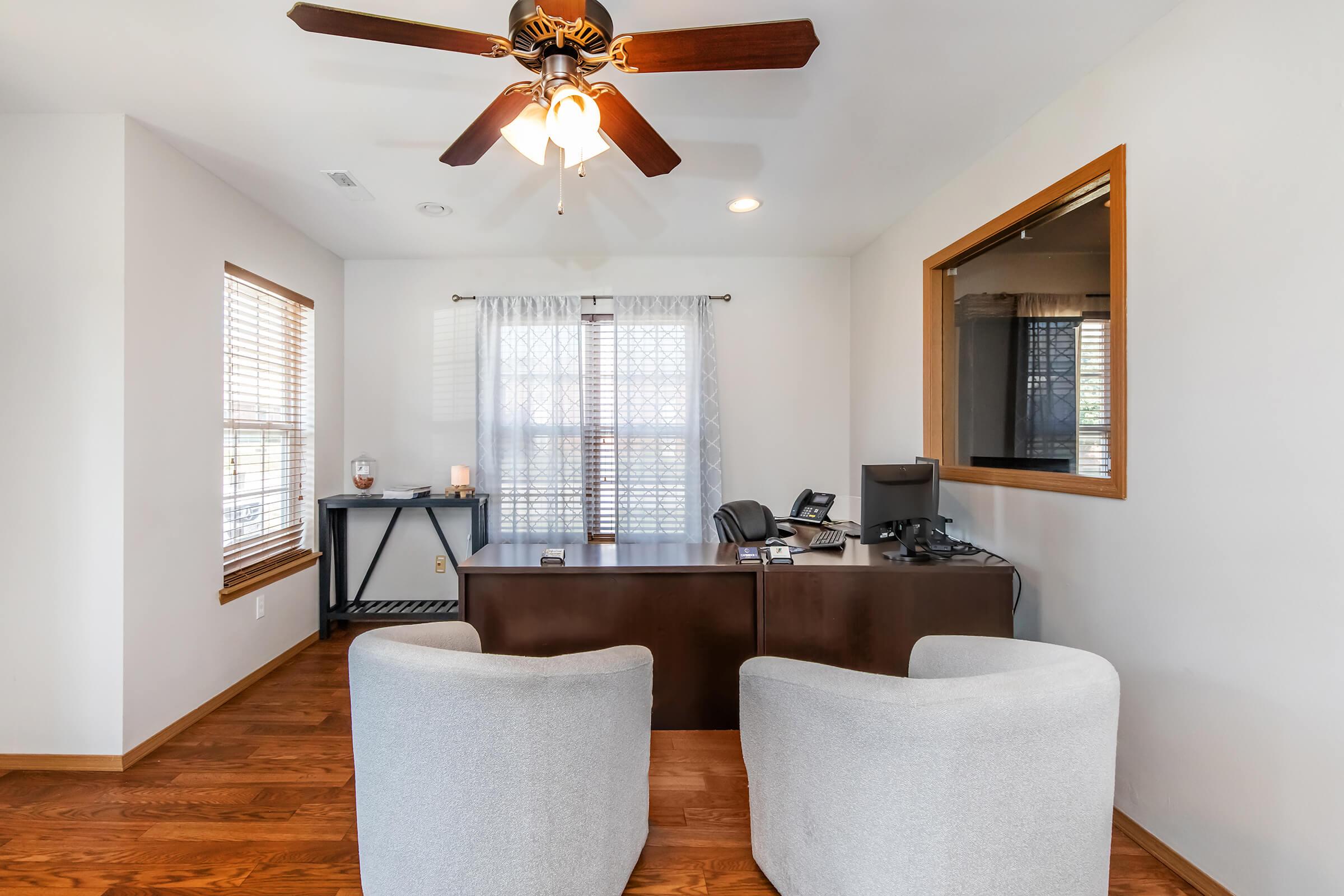 A cozy office space featuring two light gray armchairs facing a wooden desk. The desk has a computer and phone, with a window behind it covered by sheer curtains. Natural light filters in, enhancing the warm wooden floor and light walls, creating a welcoming and productive atmosphere.