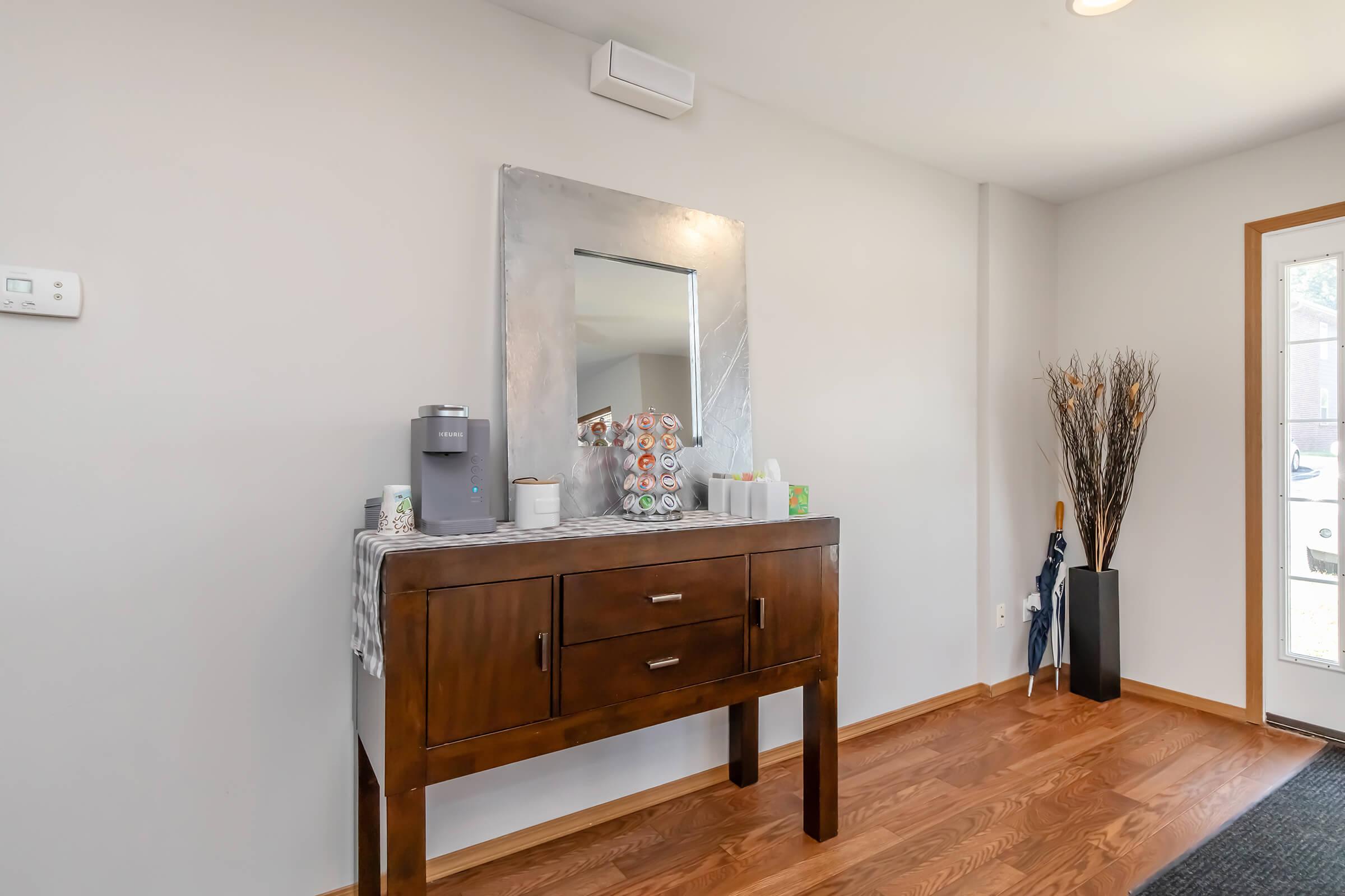 A stylish entryway featuring a wooden console table with a decorative tablecloth. On the table, there's a coffee maker and various cups. Above the table, a large mirror with a simple frame reflects the light. The wall is painted a soft color, and a tall decorative plant stands in the corner near a door.