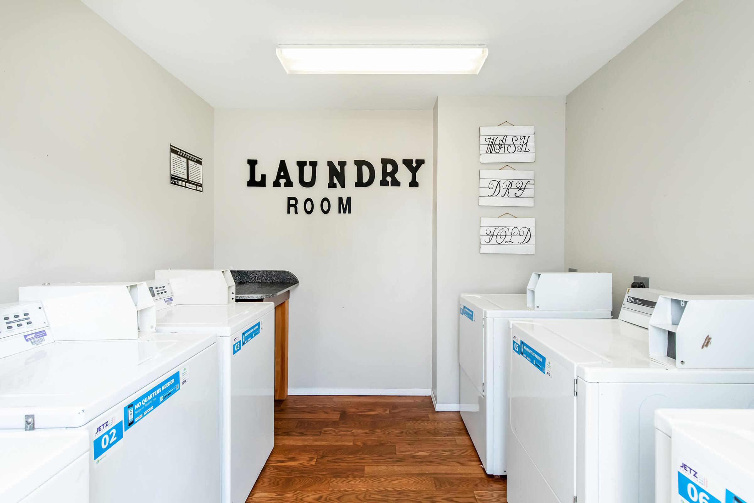 Bright laundry room featuring several white washing machines and a countertop. The wall displays the words "LAUNDRY ROOM" in bold letters, complemented by decorative wall art. The floor is made of wood, creating a clean and functional atmosphere for laundry tasks.