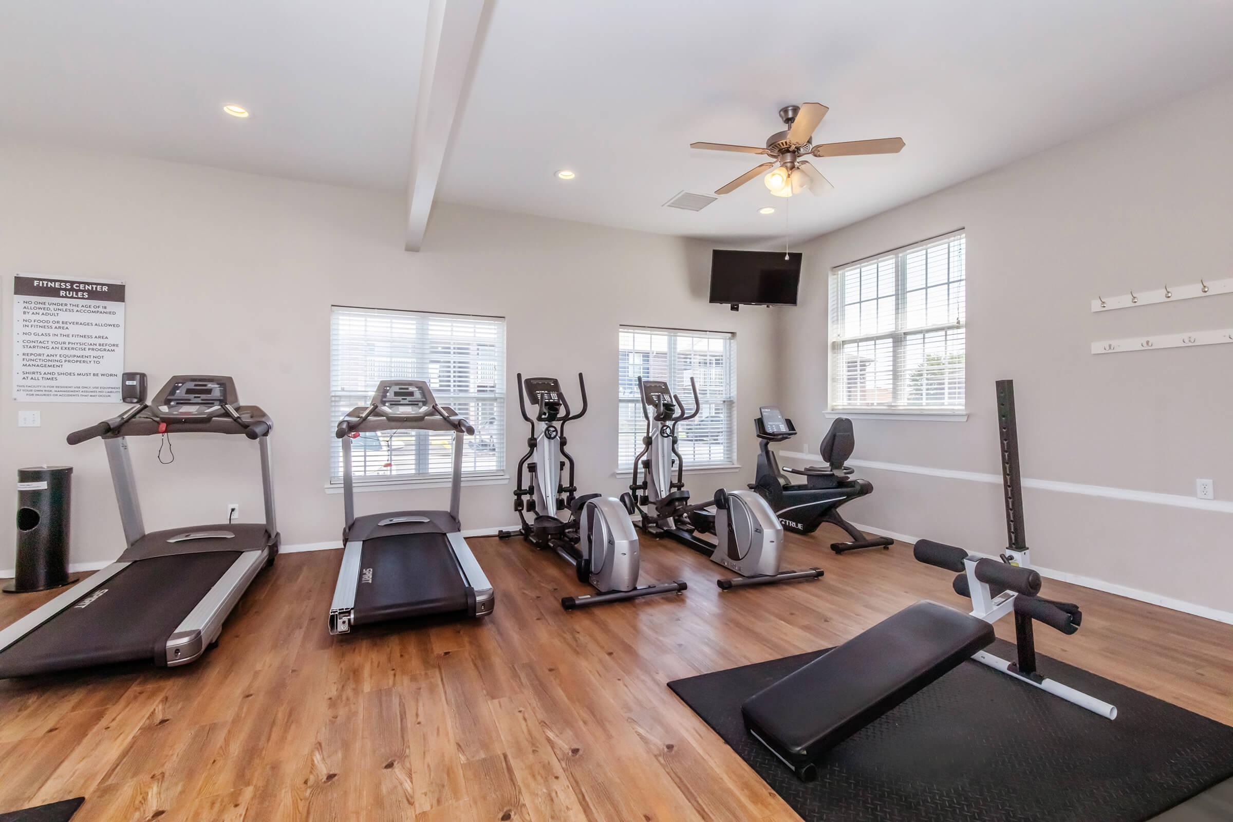 A well-lit gym interior featuring exercise equipment. There are three treadmills, a stationary bike, and a weight bench on a wooden floor. Windows allow natural light, and a ceiling fan provides ventilation. A wall-mounted TV is visible, along with a sign outlining gym rules and information.