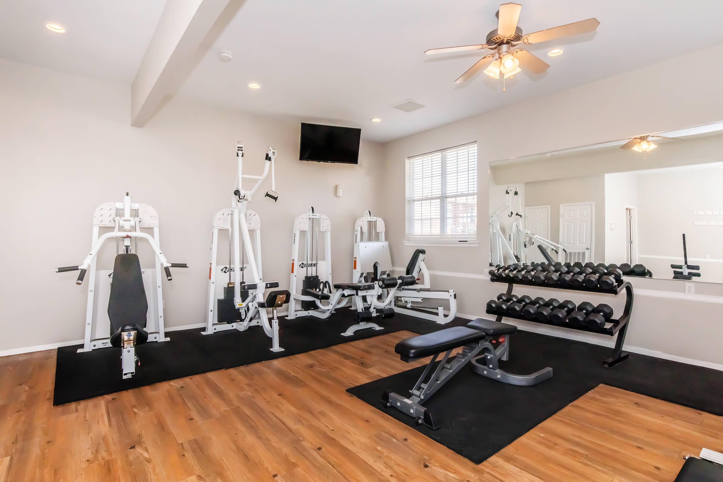 A well-equipped home gym featuring various strength training machines, a set of dumbbells on a rack, a bench, and a large mirror. Natural light enters through a window, and a ceiling fan is installed for ventilation. The flooring is wood-look laminate, creating a warm atmosphere.