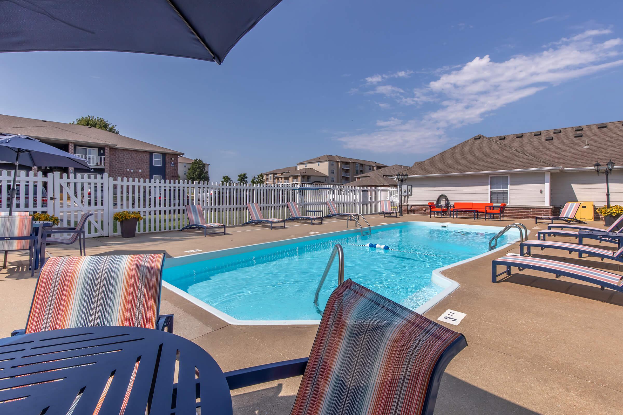 A sunny outdoor swimming pool surrounded by lounge chairs and tables. An umbrella provides shade, while nearby apartment buildings and a white picket fence complete the scenic view. Clear blue skies reflect over the water, creating a relaxing atmosphere.
