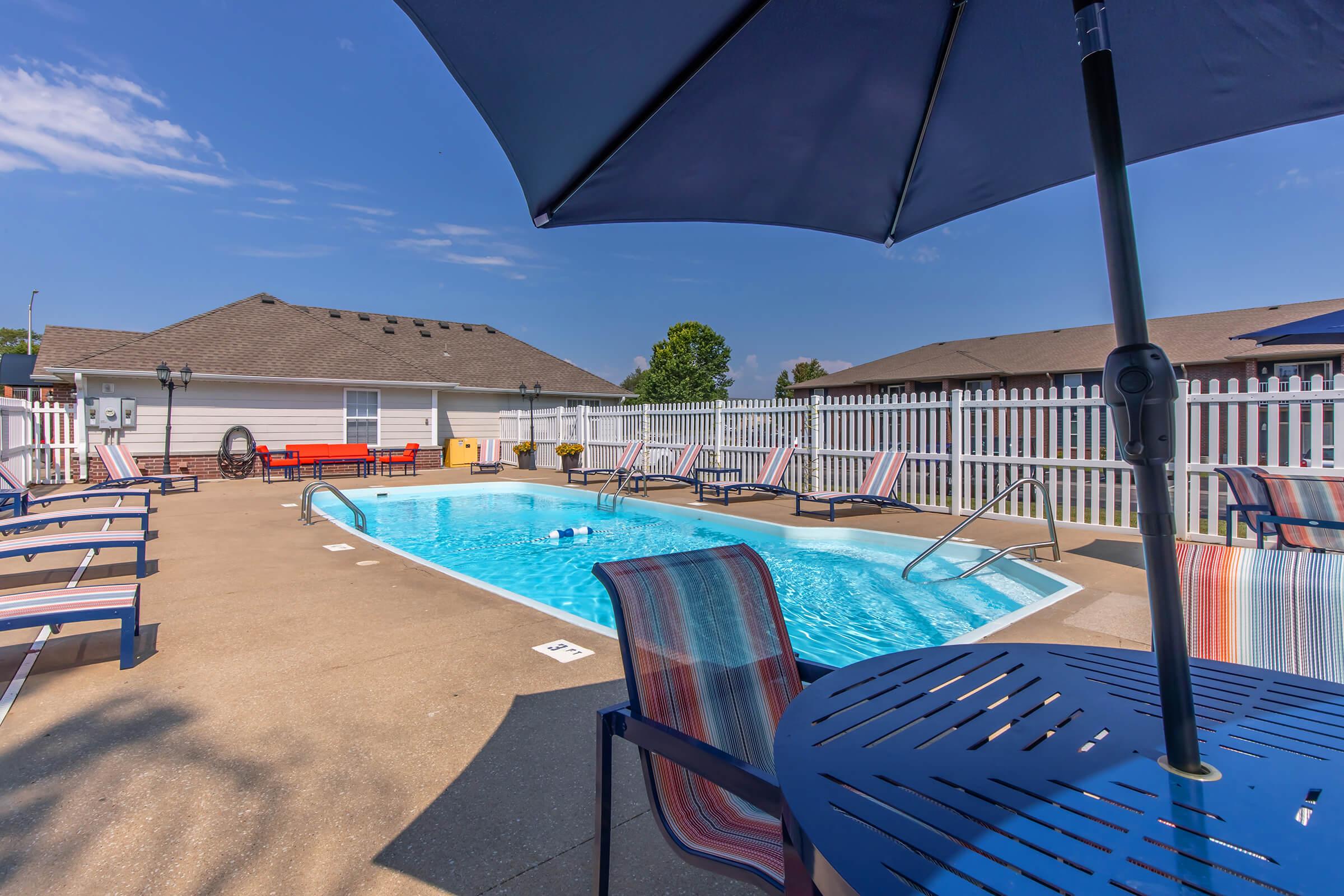 A serene outdoor swimming pool area featuring a clear blue pool surrounded by lounge chairs and tables. An umbrella provides shade over a table, and a white picket fence encloses the space. The sky is clear with a few clouds, creating a relaxing atmosphere perfect for leisure and enjoyment.