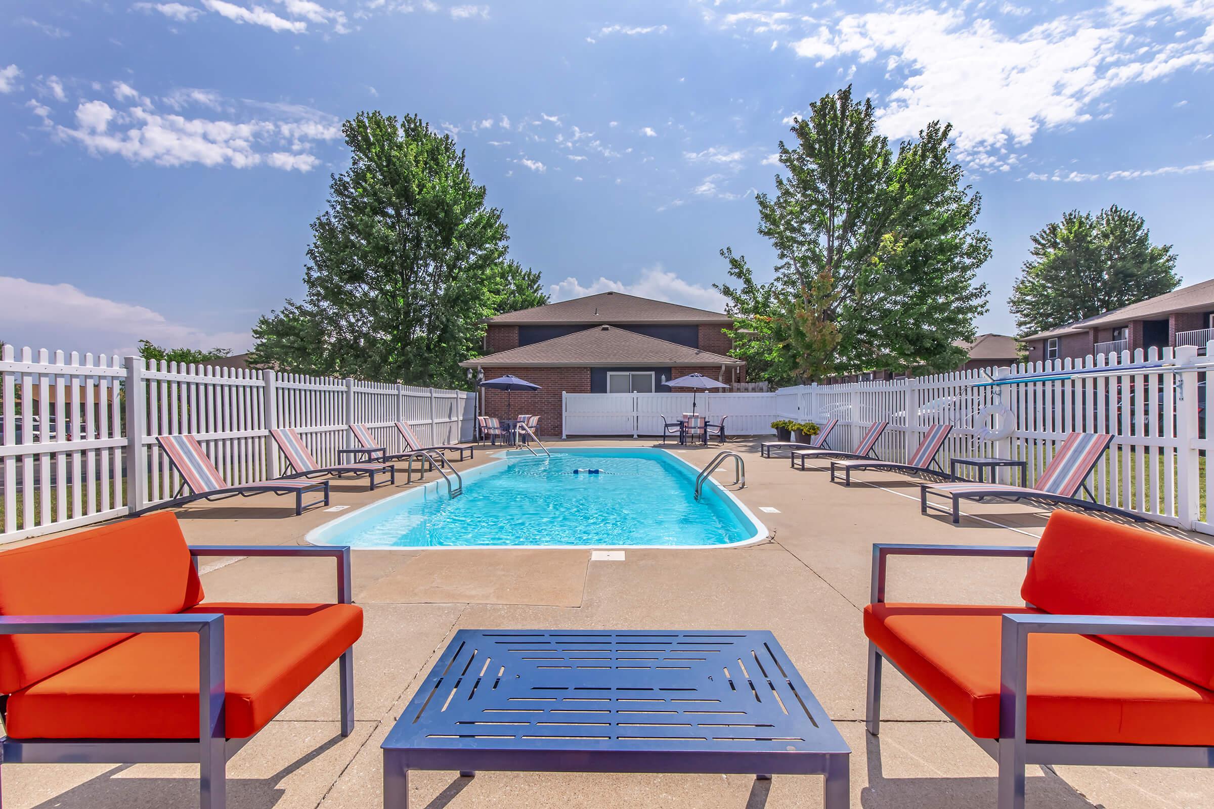 A view of a swimming pool surrounded by a white fence, with lounge chairs arranged around it. Orange outdoor chairs and a coffee table are in the foreground. Bright blue sky with scattered clouds and trees in the background add to the inviting atmosphere of the pool area.