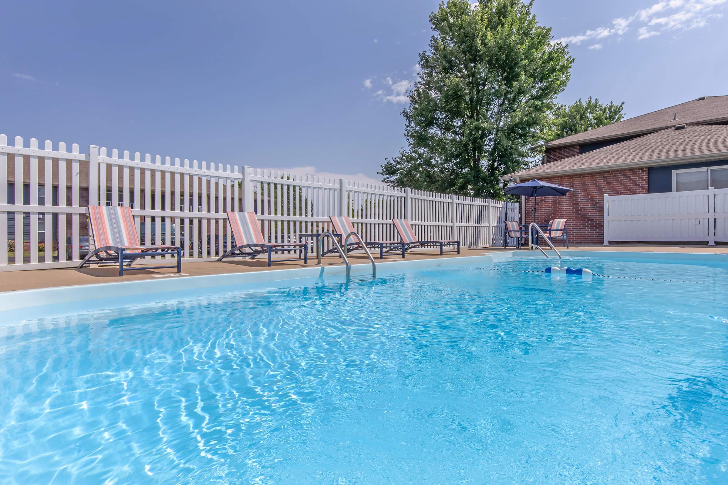 A clear blue swimming pool surrounded by a paved area, featuring several lounge chairs with striped cushions. A patio umbrella provides shade, and a white picket fence encloses the area. The sky is bright with a few clouds, and a tree is visible in the background.