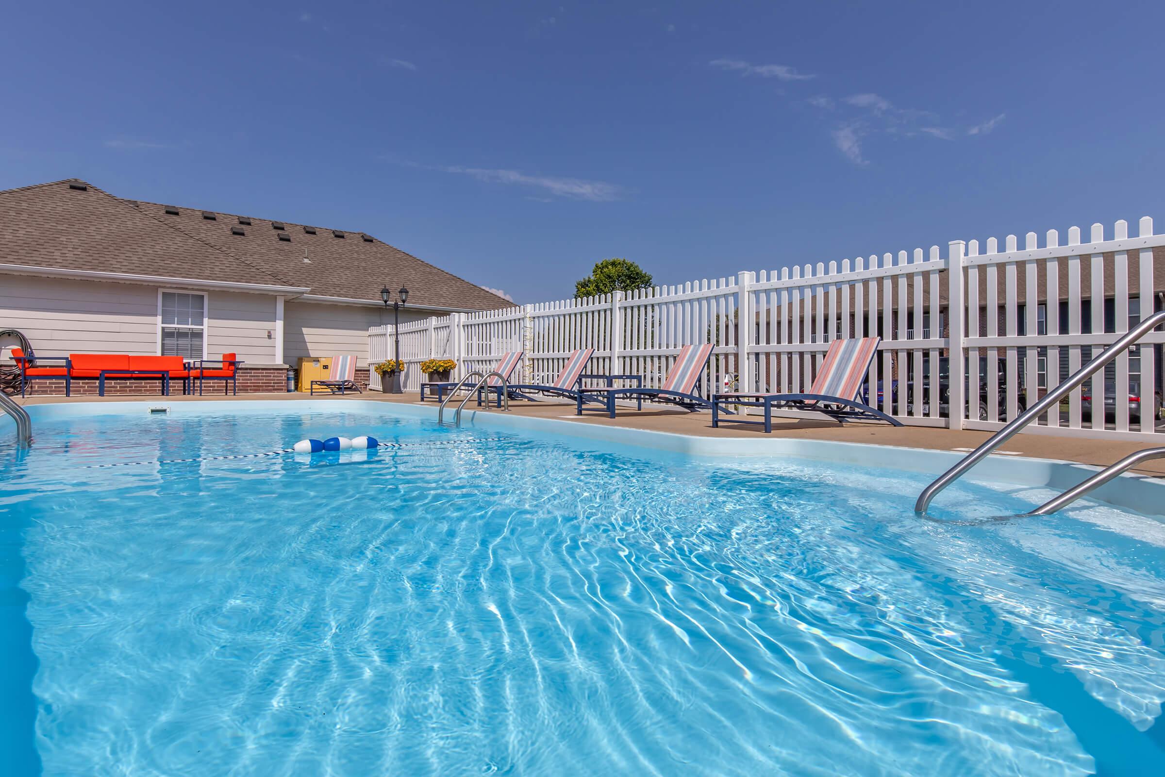 A clear blue swimming pool with a floating pool toy, surrounded by lounge chairs. In the background, a white picket fence and a building with a rooftop are visible under a sunny sky. The scene conveys a peaceful and inviting atmosphere for relaxation and leisure.