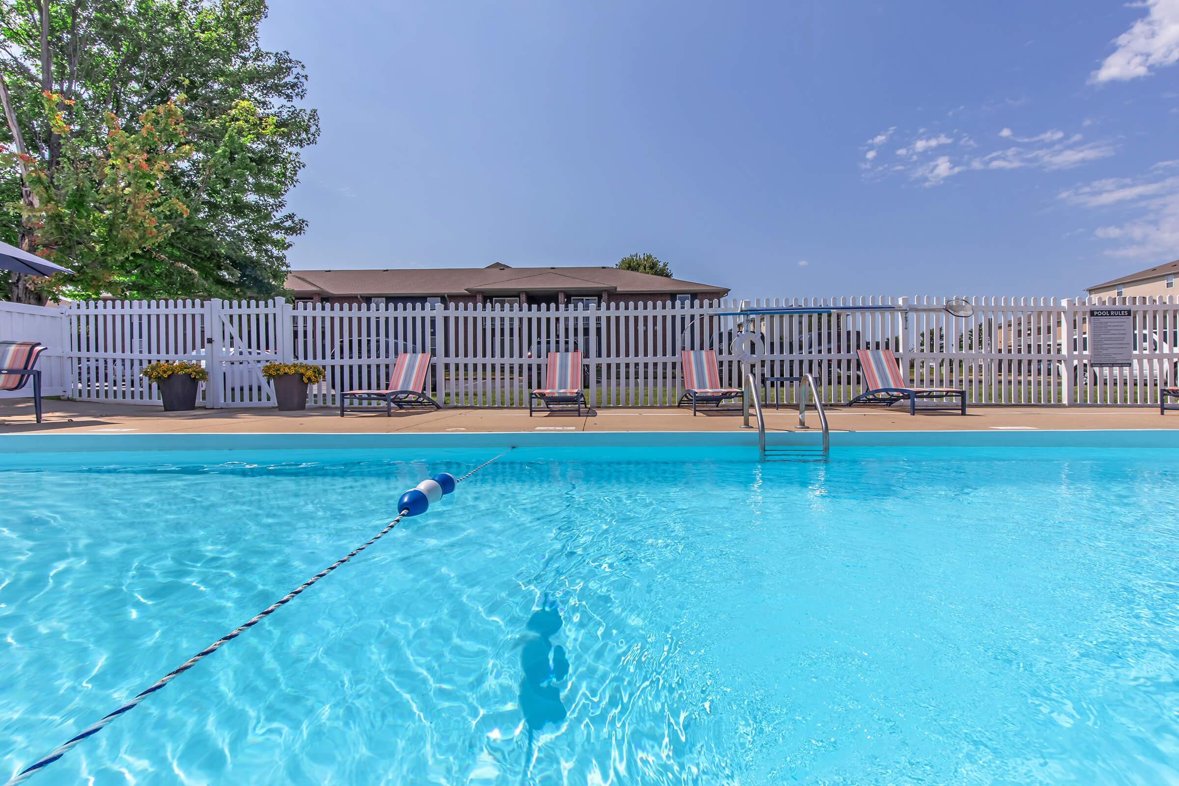 A clear blue swimming pool surrounded by lounge chairs and a white picket fence. Green trees are visible in the background under a bright blue sky with a few clouds. The pool has a diving board and a lane marker, creating a serene and inviting atmosphere for relaxation.