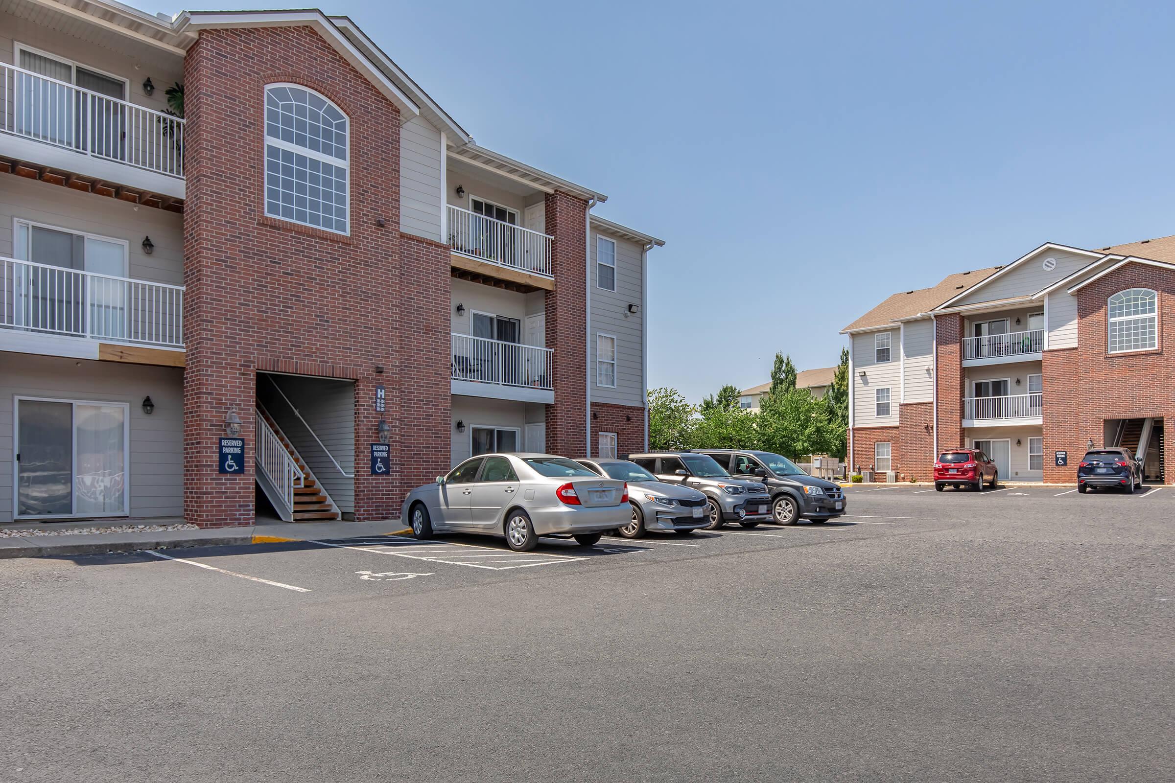Residential apartment complex with two buildings. The building in the foreground features a red brick facade, balconies, and large windows. A parking lot is visible in front, occupied by several cars. The sky is clear and bright, indicating a sunny day.