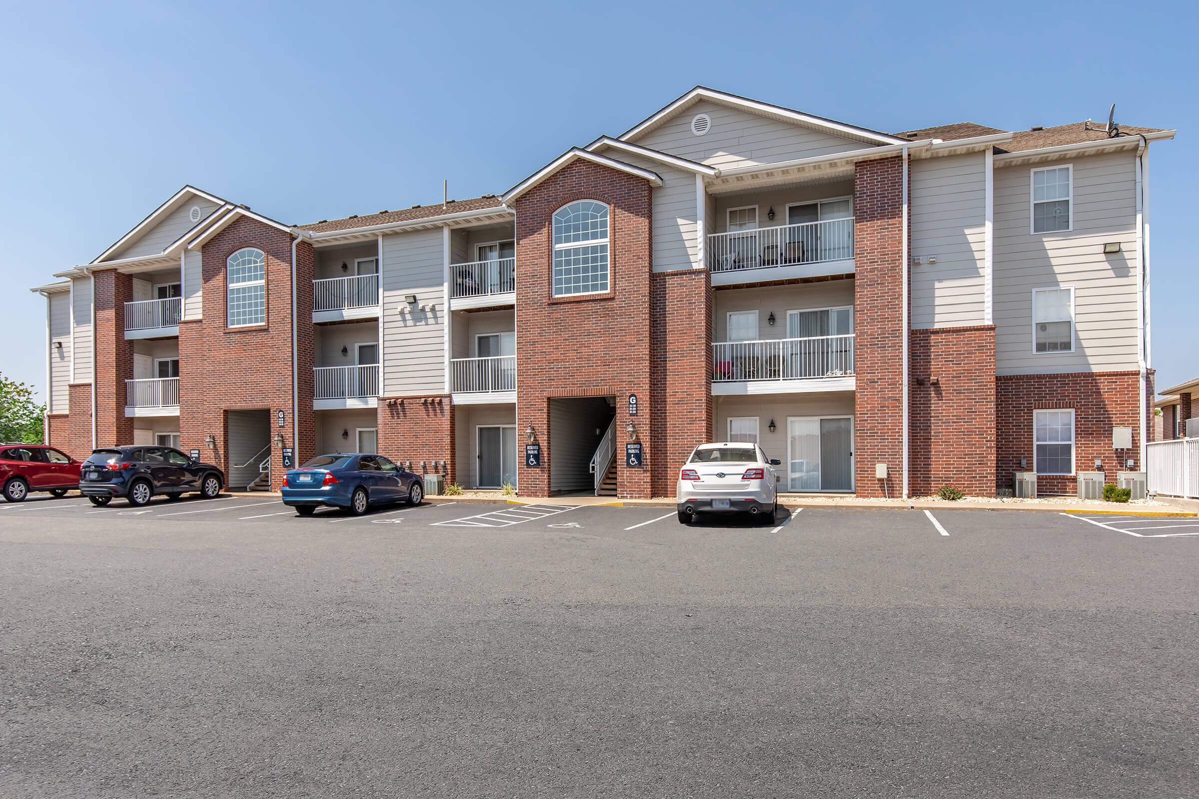 A three-story brick and siding apartment building with multiple balconies. It features a parking lot with several cars parked in front. The sky is clear and blue, and there are no visible people in the image.