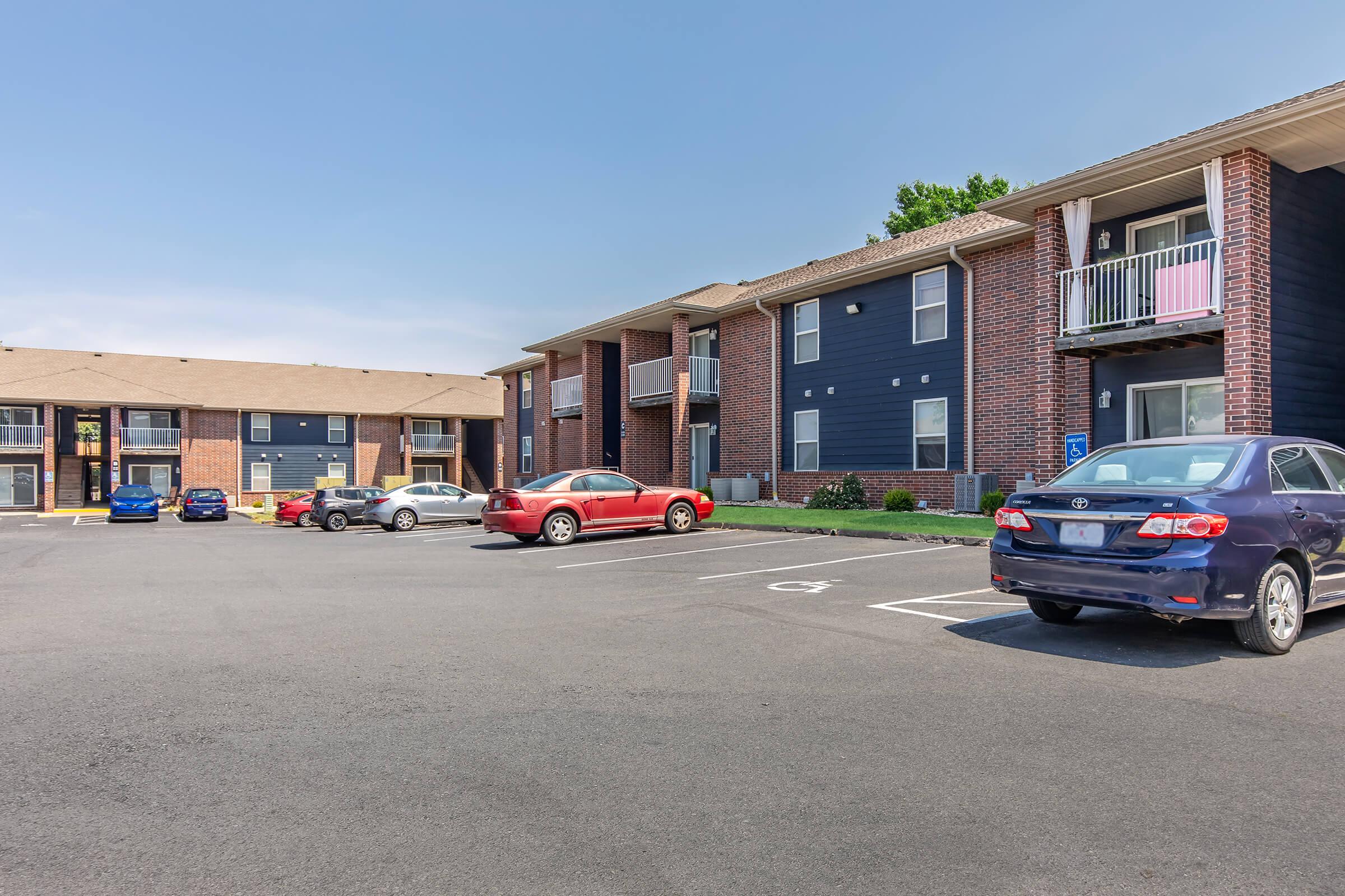A parking lot outside a two-story apartment complex. The building has a mix of brick and blue siding, with several cars parked in front. Balconies are visible on the upper floor, and the sky is clear and bright. The area appears well-maintained and inviting.