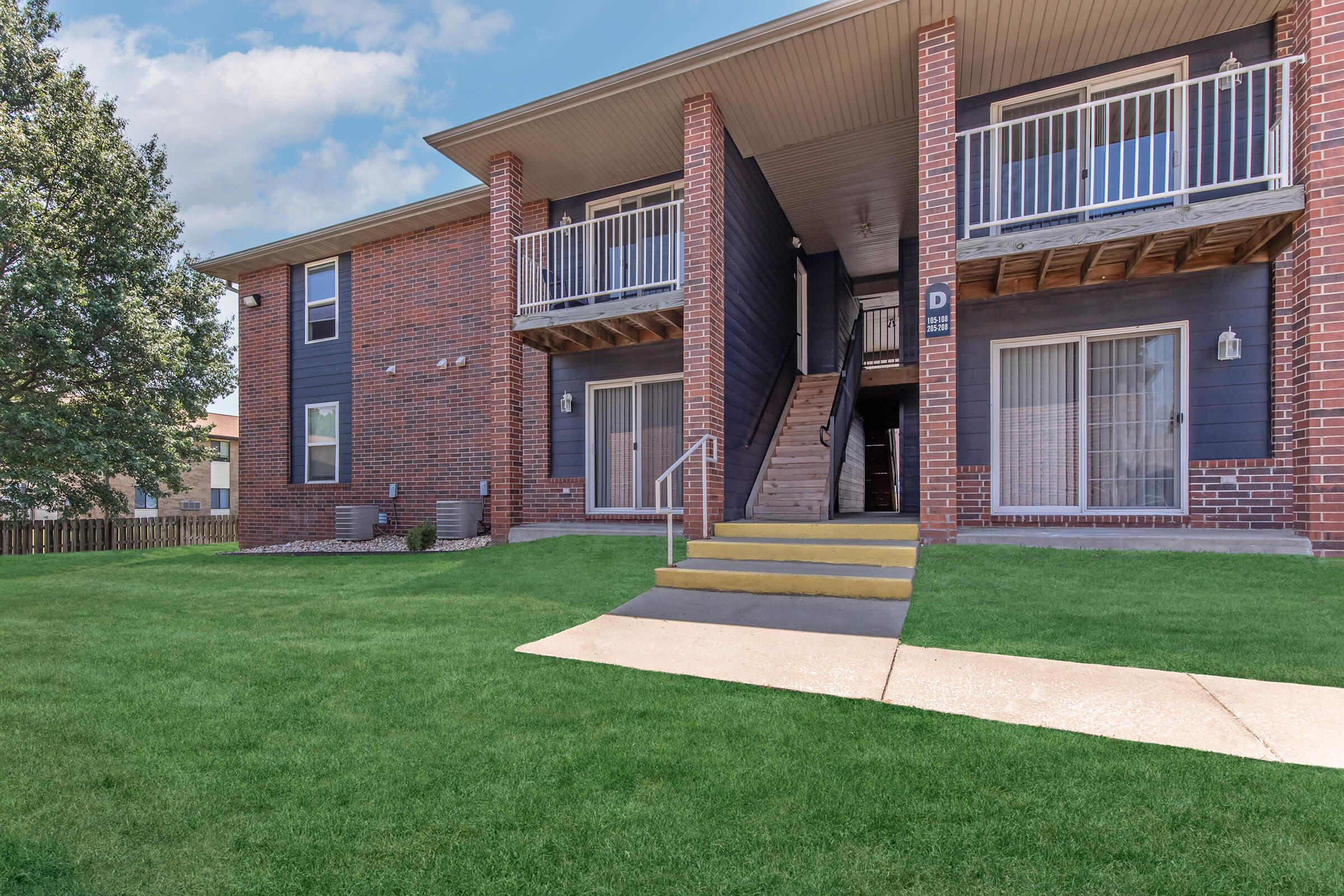 A multi-unit residential building with brick and blue siding. The foreground features a well-maintained lawn and a concrete pathway leading to a set of stairs that access the upper units. The exterior includes balconies and large windows, highlighting a welcoming and modern architectural style.