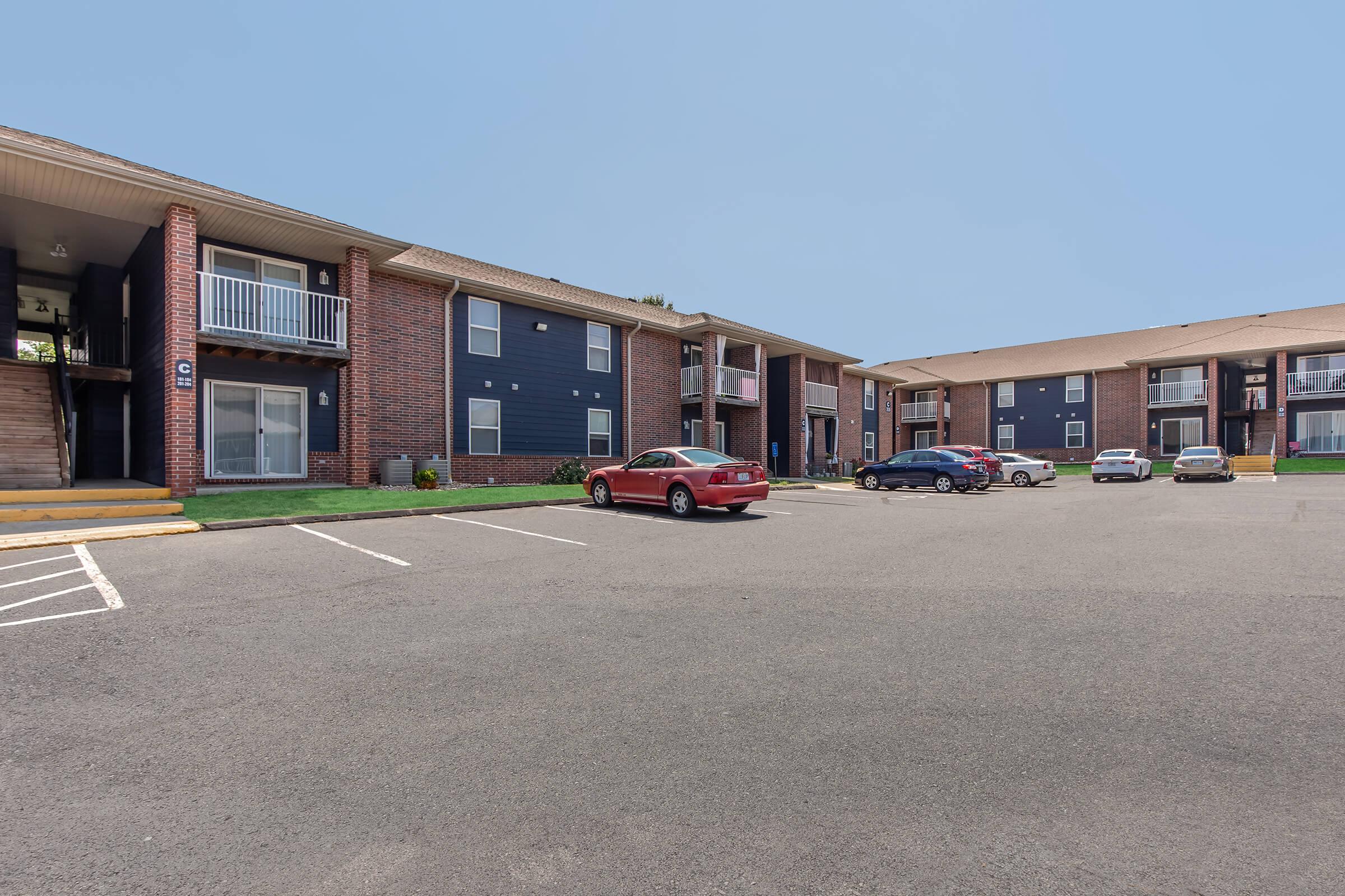 View of a residential apartment complex featuring two-story brick buildings with balconies. A parking lot is visible in the foreground with several parked cars. The sky is clear and blue, and the surrounding area is well-maintained with green grass.