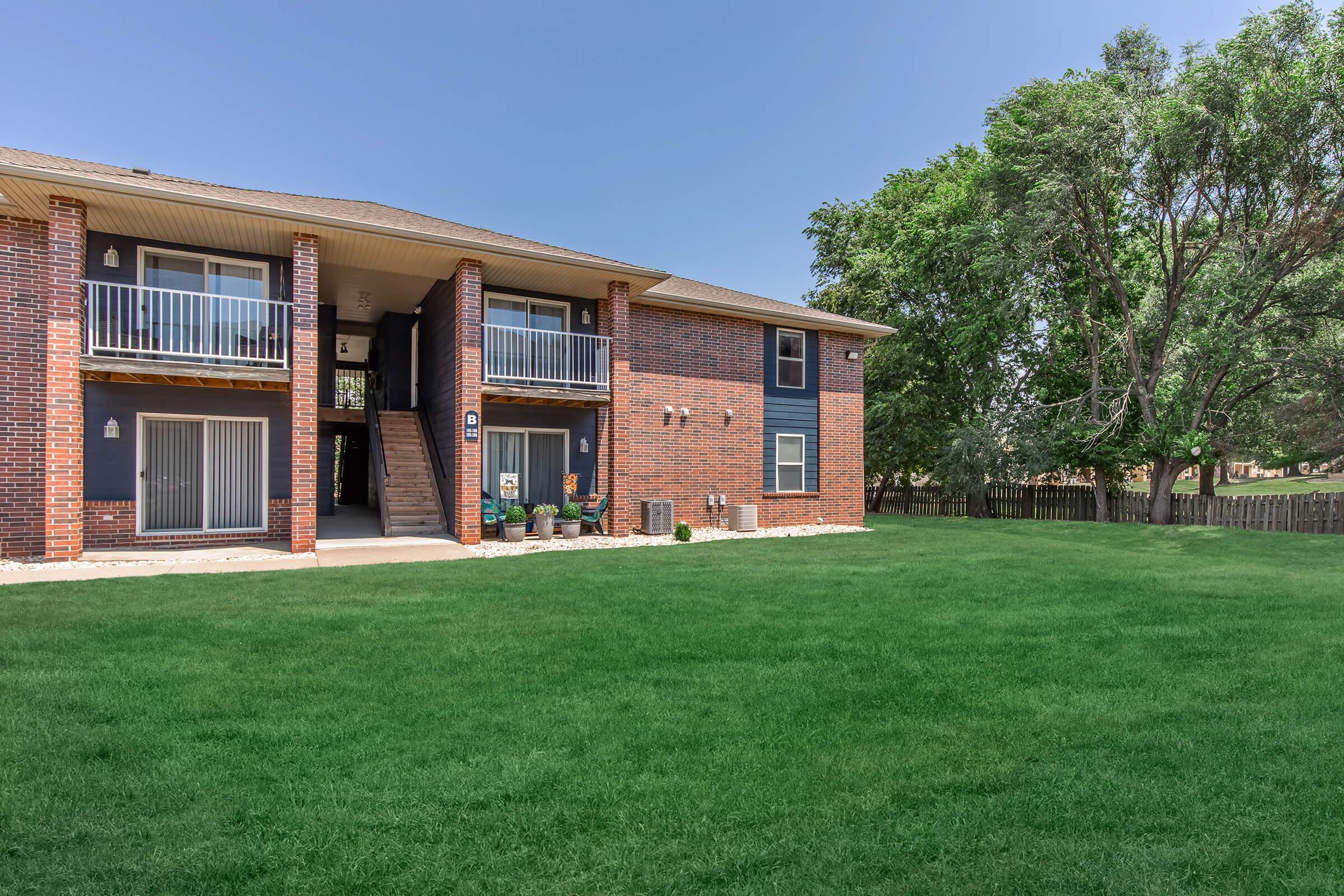 Two-story brick apartment building with a grassy lawn, featuring balconies on the upper floors. Trees are visible to the right, and the exterior shows a well-maintained area with steps leading to the entrance. The sky is clear and blue.