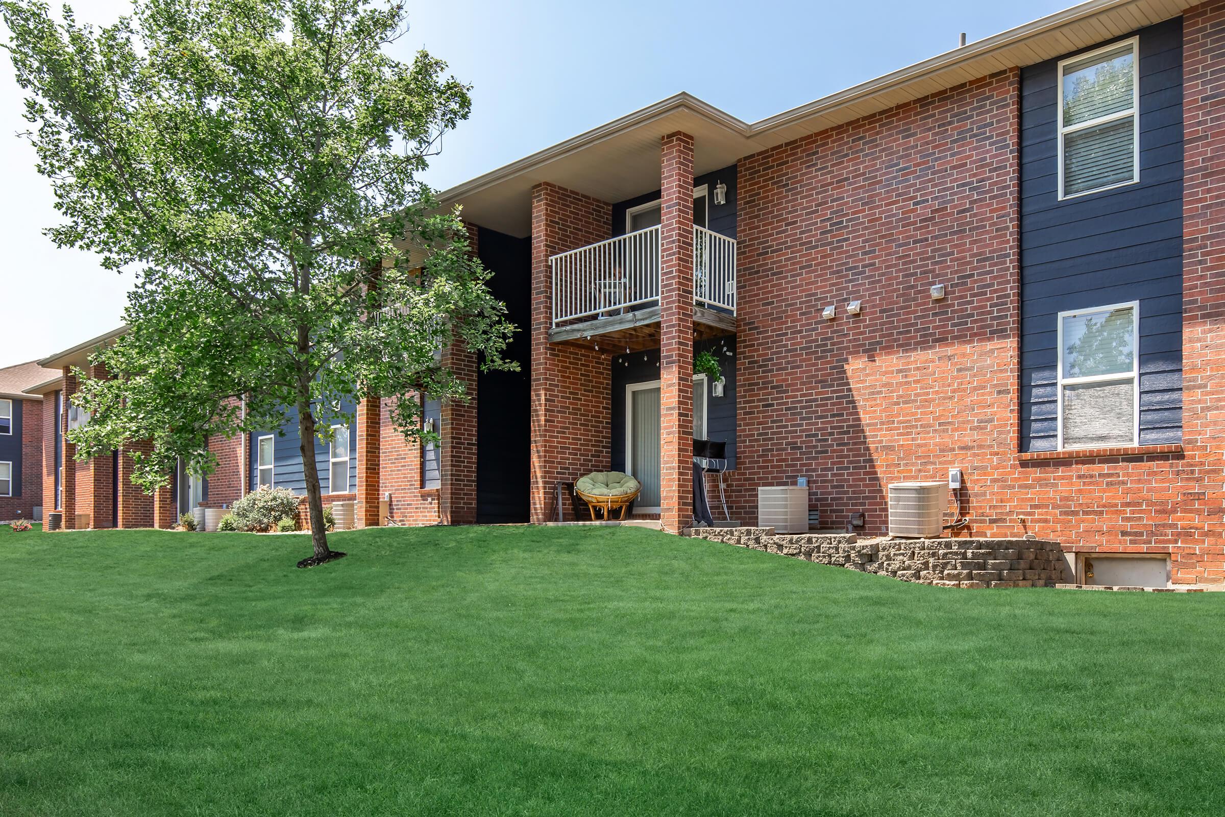 A well-maintained apartment building featuring brick and dark siding, with a green lawn in front. A small tree adds to the landscaping. The entrance includes a balcony, and there are two air conditioning units visible on the ground level.