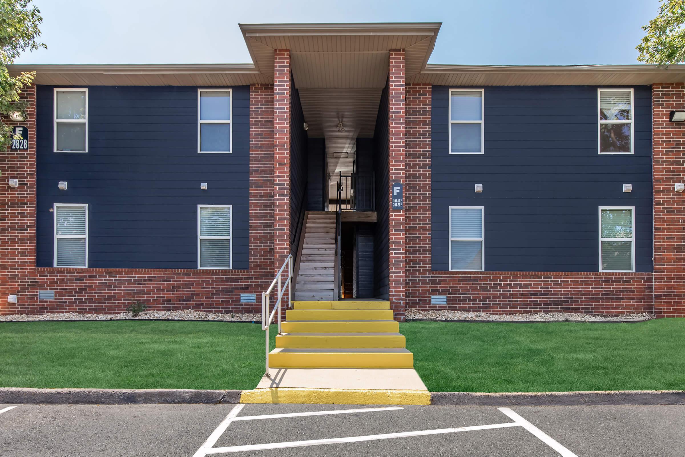 Two-story apartment building with a brick exterior and dark siding. The entrance features a set of yellow stairs leading to a door, with two windows visible on each side of the staircase. The surrounding area includes manicured grass and a parking lot below.
