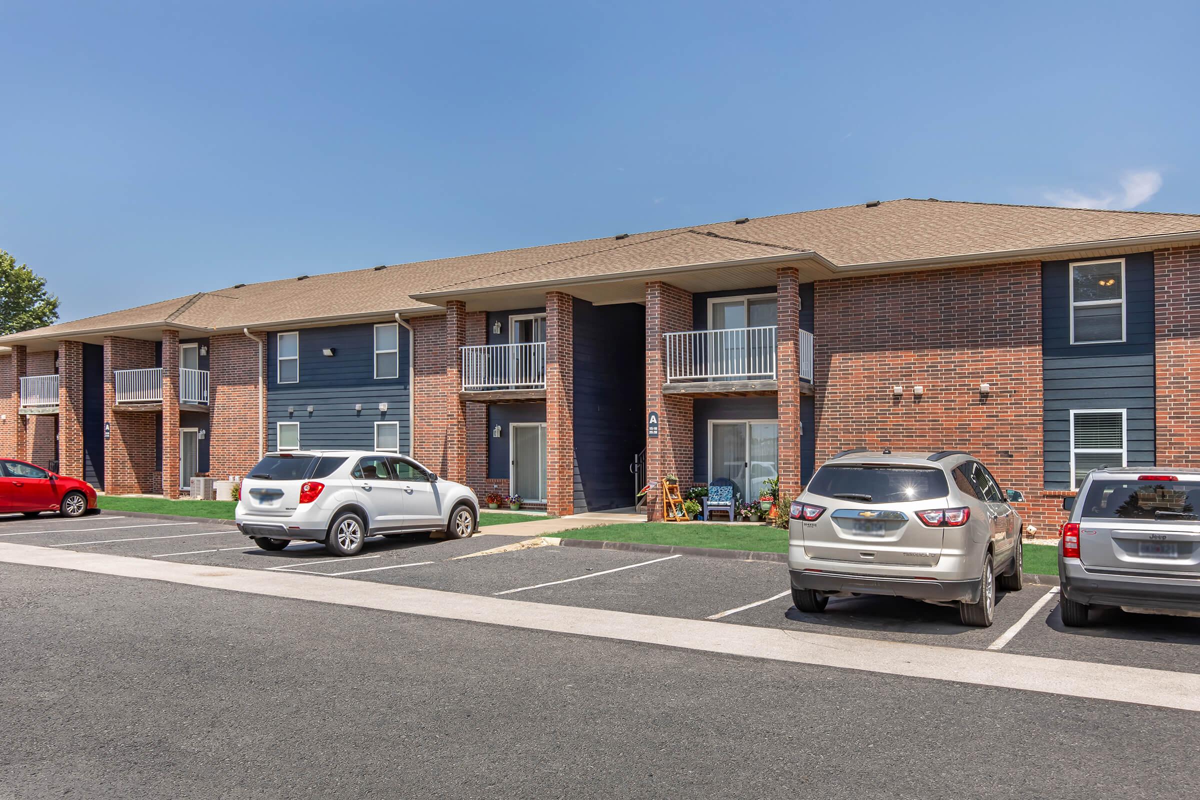 Two-story apartment building with brick and dark blue siding. Balconies are visible on the upper floor. Several cars parked in front, including a red sedan, a white SUV, and a silver sedan. Lawn area in front of the building with a clear blue sky above.