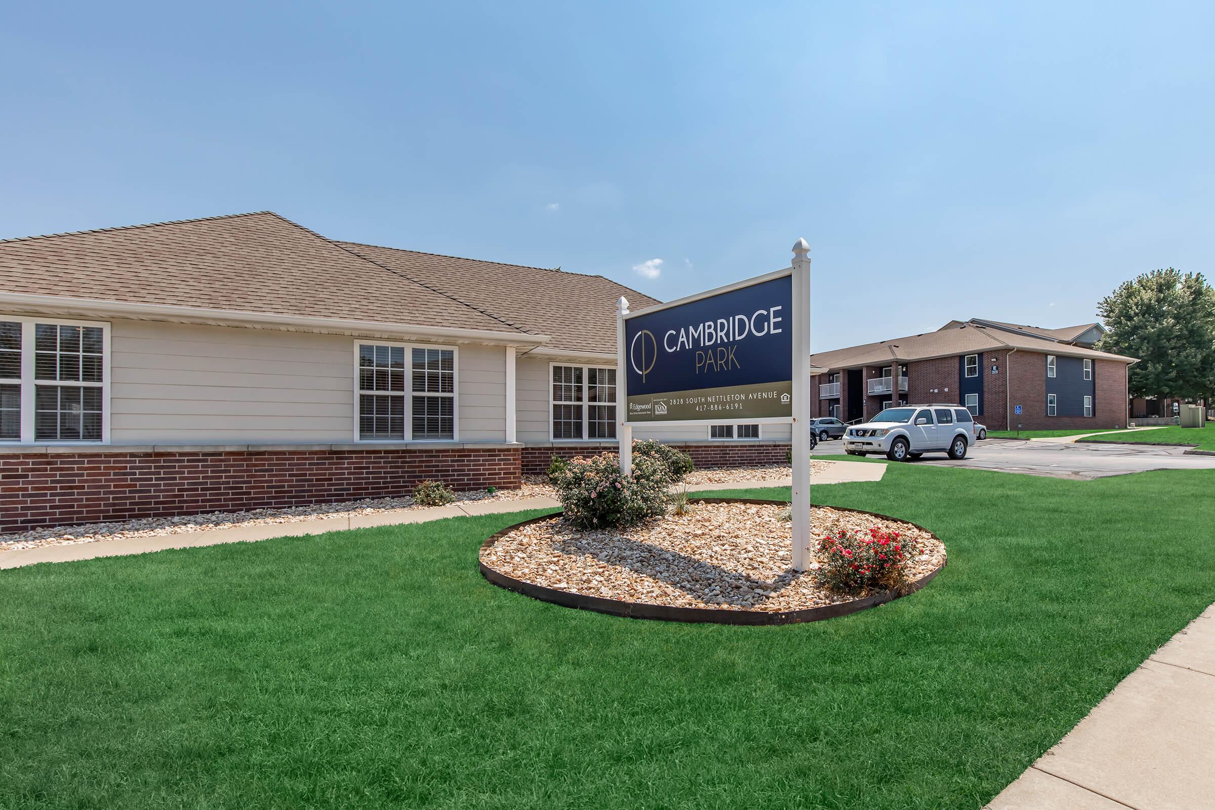 A sign for Cambridge Park is displayed in front of a well-maintained building. The landscape features green grass and flower beds, while a residential building can be seen in the background. The sky is clear, indicating a sunny day.