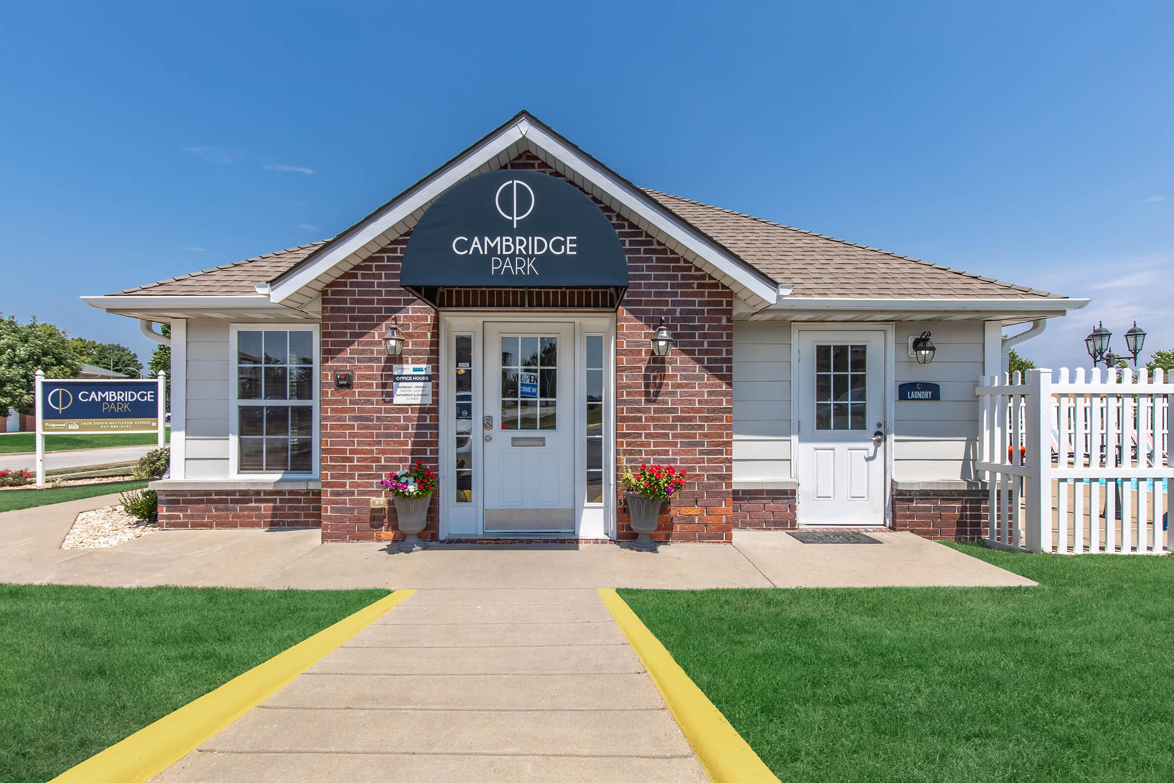 A brick building with a sloped roof and a sign reading "Cambridge Park." The entrance features a door with a window, surrounded by flower pots. There’s a white door to the right, and a well-maintained lawn leads up to the entrance. A blue sky is visible in the background.