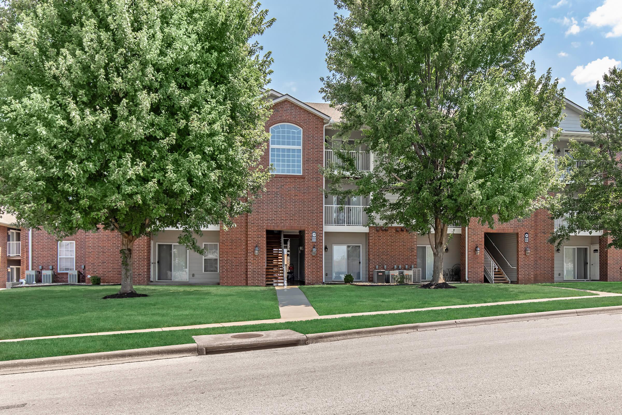 A brick apartment building with large windows and two symmetrical trees in front. The building features multiple entrances, a green lawn, and a nearby sidewalk. The sky is clear with a few clouds, and the scene conveys a well-maintained residential area.