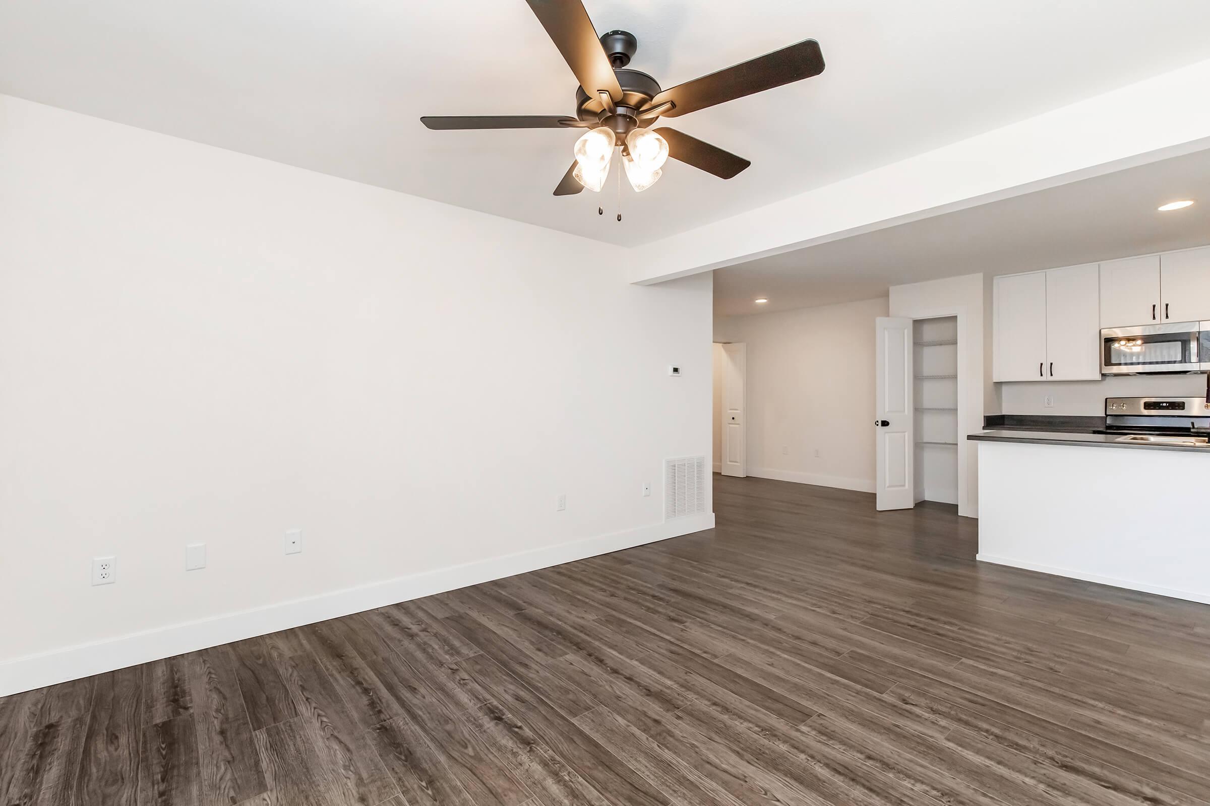 Spacious, modern living area featuring a ceiling fan, wood-like flooring, and an open layout connected to a kitchen with stainless steel appliances. The walls are painted white, contributing to a bright and airy atmosphere. A doorway leads to additional spaces, enhancing functionality.