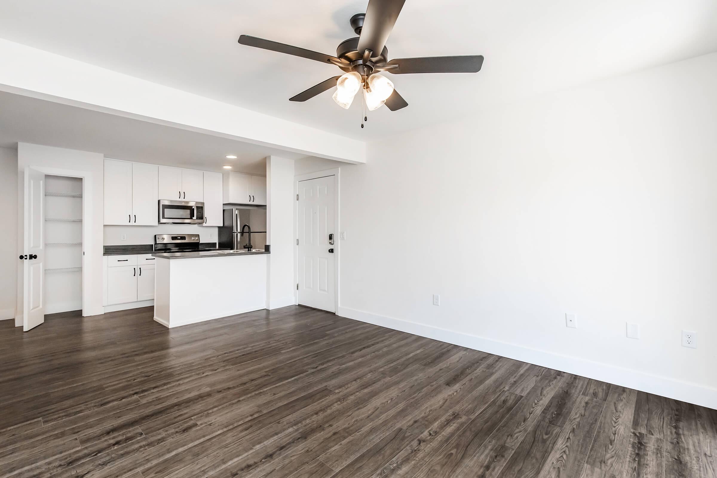 Modern apartment interior featuring an open floor plan. The living area has hardwood-style flooring and a ceiling fan, while the kitchen is visible in the background with white cabinets and stainless steel appliances. Bright, neutral walls enhance the airy feel of the space.