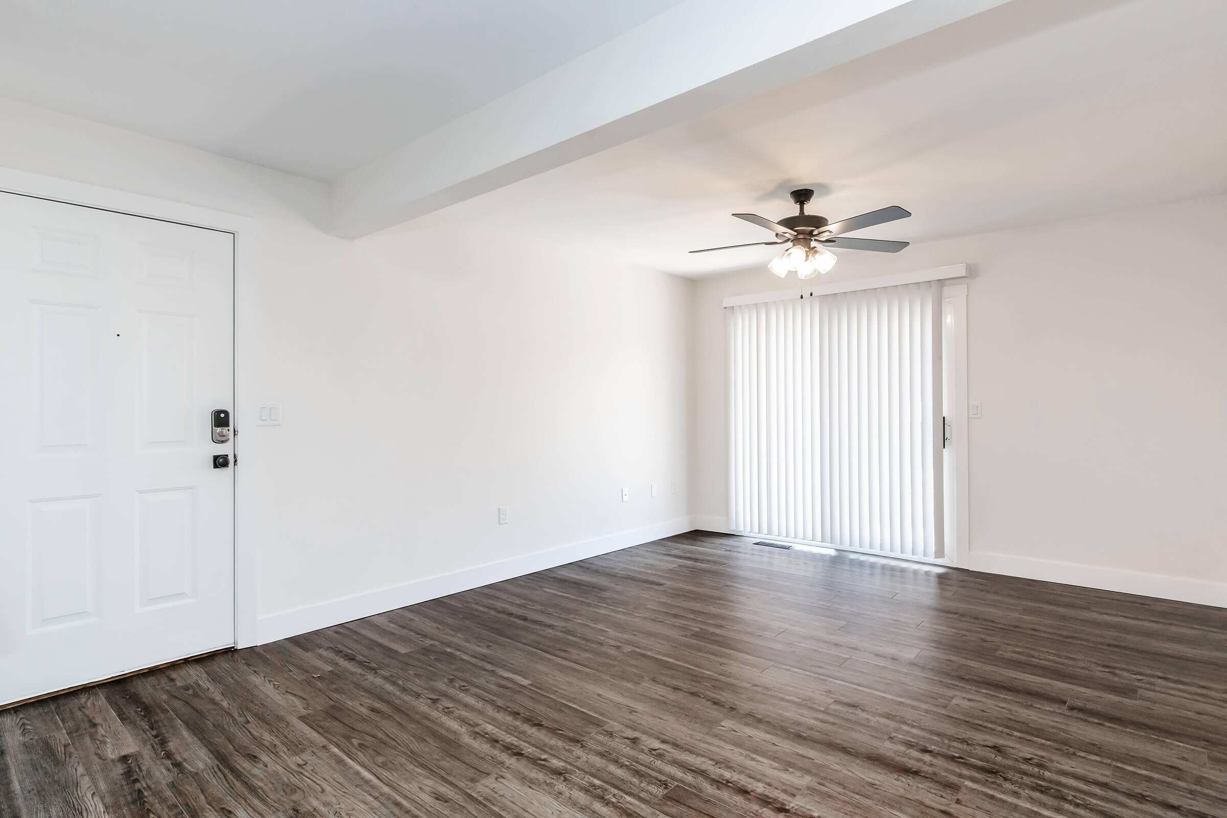 A bright, empty room with light-colored walls and dark wooden flooring. There is a white front door on the left, a ceiling fan in the center, and a large sliding glass door with vertical blinds on the right, leading to an outdoor space. Natural light fills the room, creating an open and airy feel.