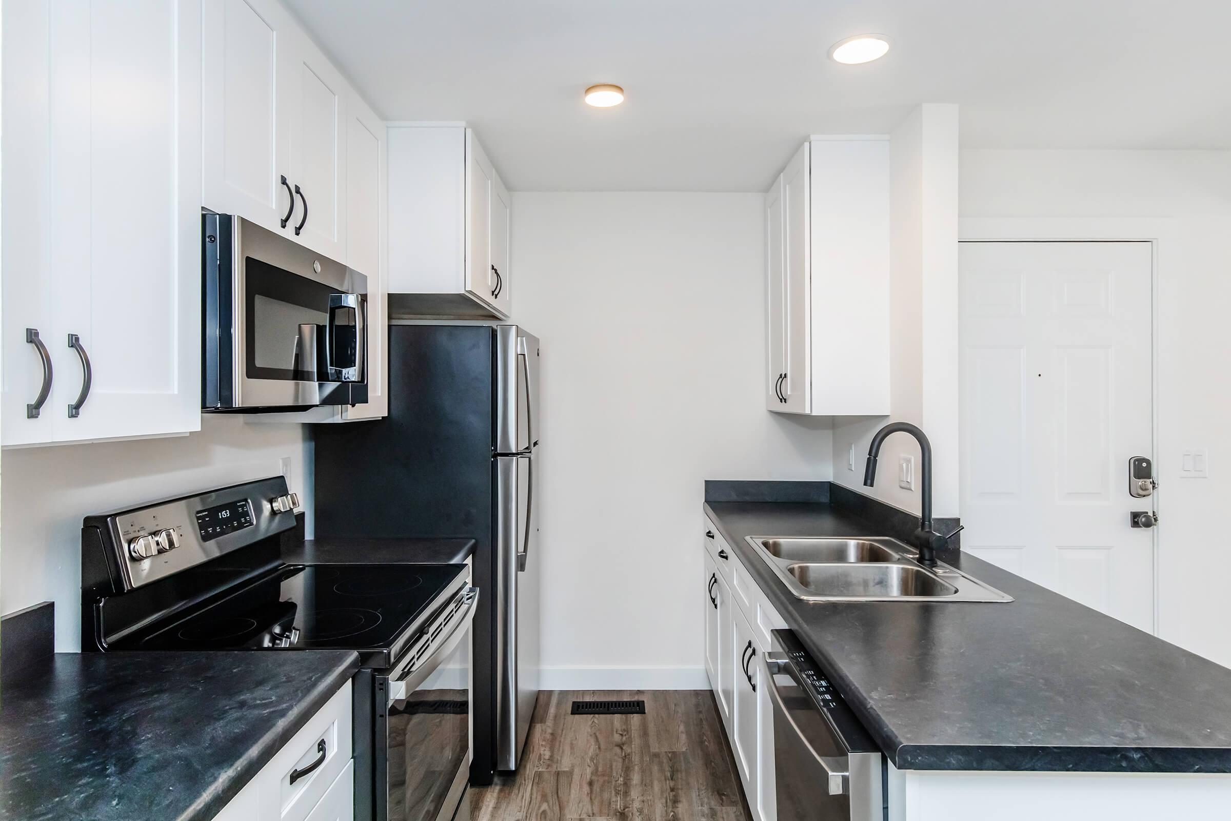 Modern kitchen featuring white cabinetry, stainless steel appliances including a microwave, refrigerator, and oven. The countertops are dark and sleek, complementing the white cabinets. Bright lights illuminate the space, which has a clean, minimalist design. A door is visible in the background.