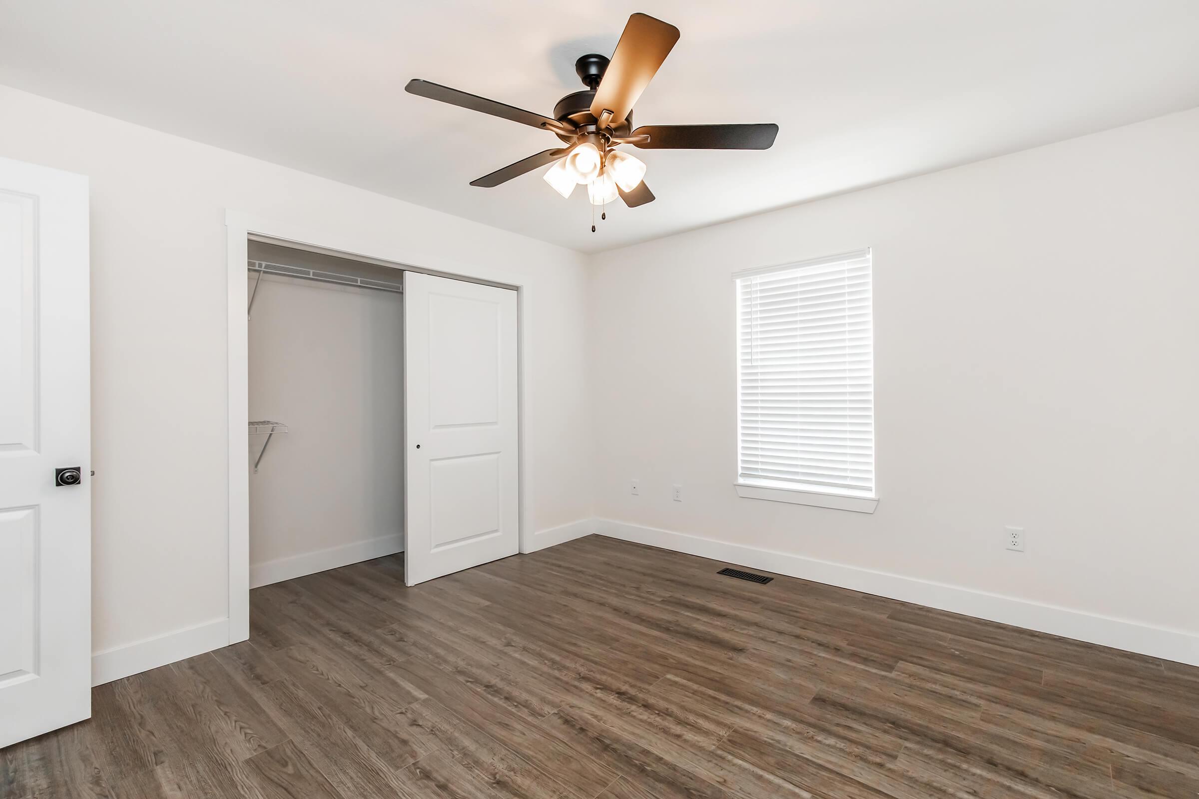 A bright and empty room featuring light-colored walls, a ceiling fan with lights, and wooden flooring. On one side, there is a closet with an open door, and a window with blinds allows natural light to enter.