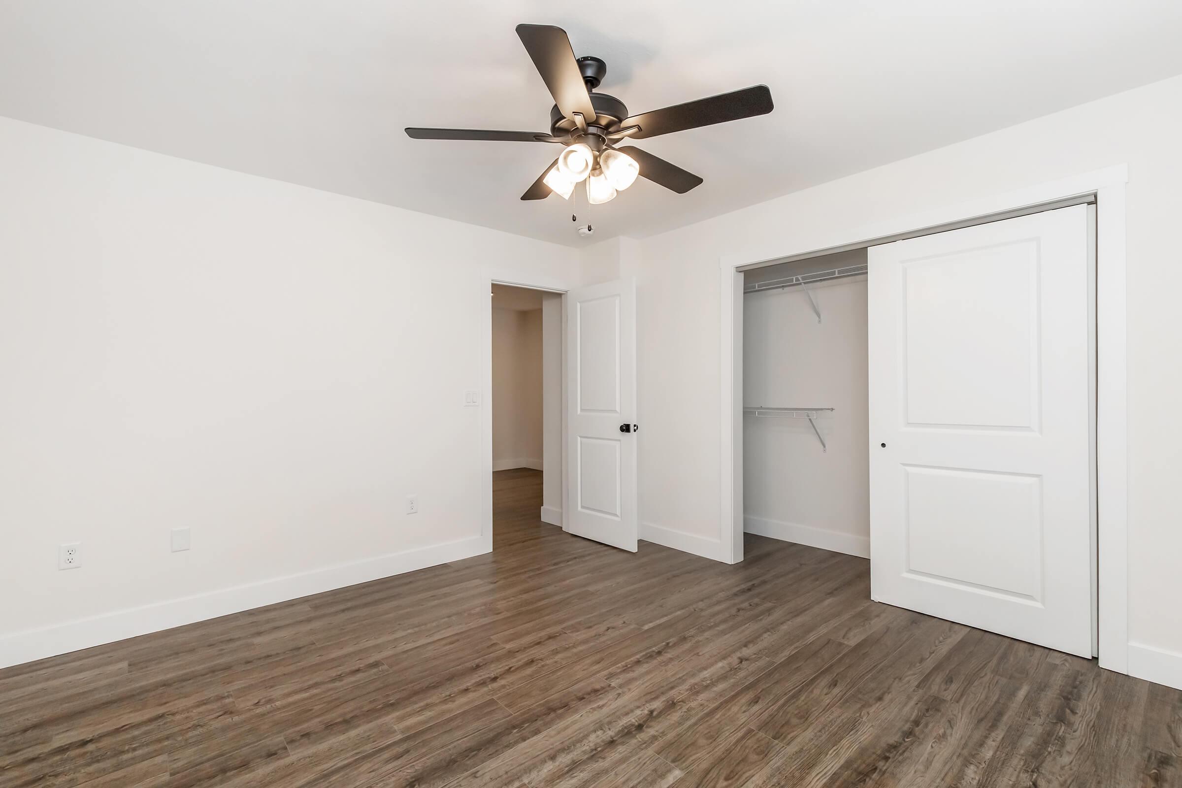 A spacious, empty room with light-colored walls and a ceiling fan featuring four lights. The room has laminate flooring and two white doors: one leading to a closet and the other to another room. The area is well-lit, showcasing a clean and modern design.