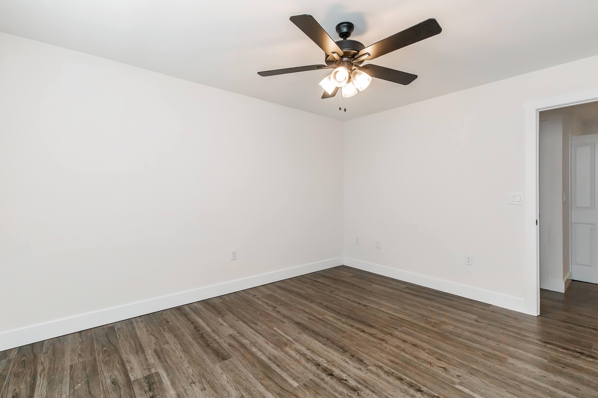 Empty room featuring light-colored walls, a ceiling fan with three bulbs, and hardwood-like flooring. There's a door on the right side leading to another room, and the space is well-lit and minimalistic, suitable for various furniture arrangements.