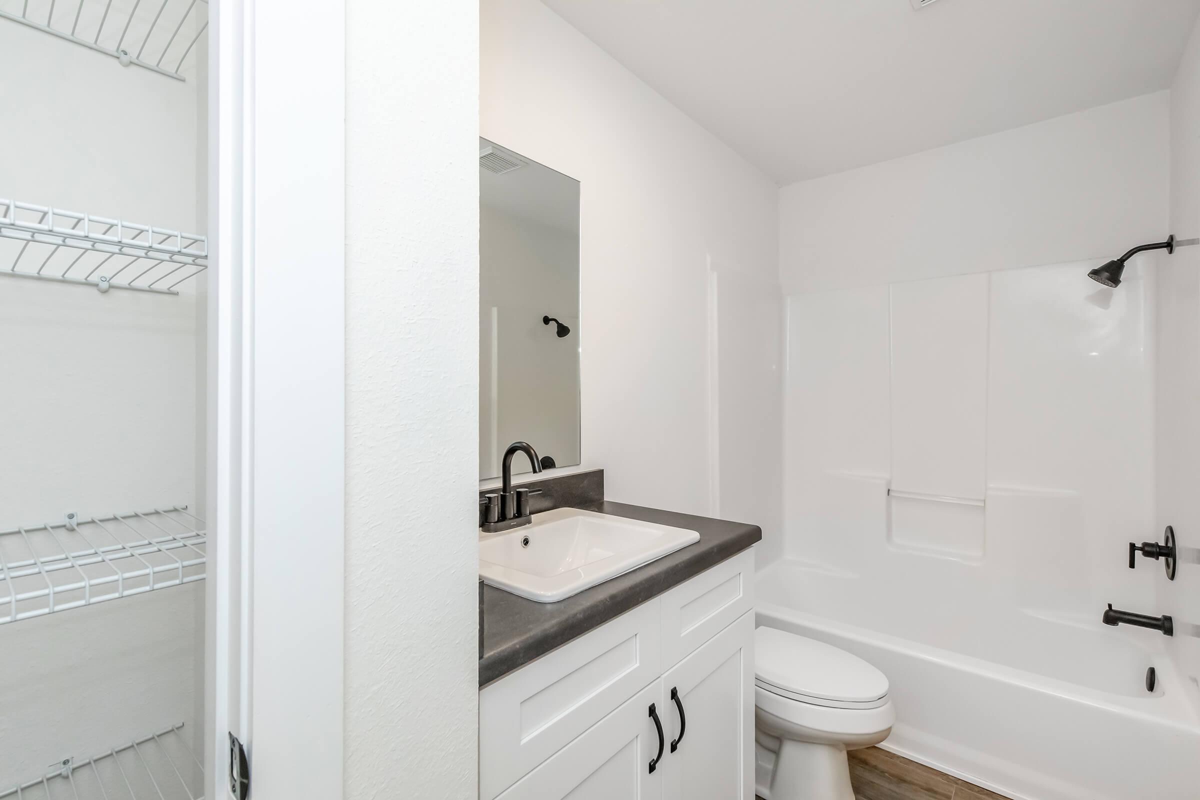 A clean, modern bathroom featuring a white bathtub, a black faucet, and a sink with a dark countertop. The walls are painted white, and there's a large mirror above the sink. To the side, an empty shelf unit provides additional storage space.