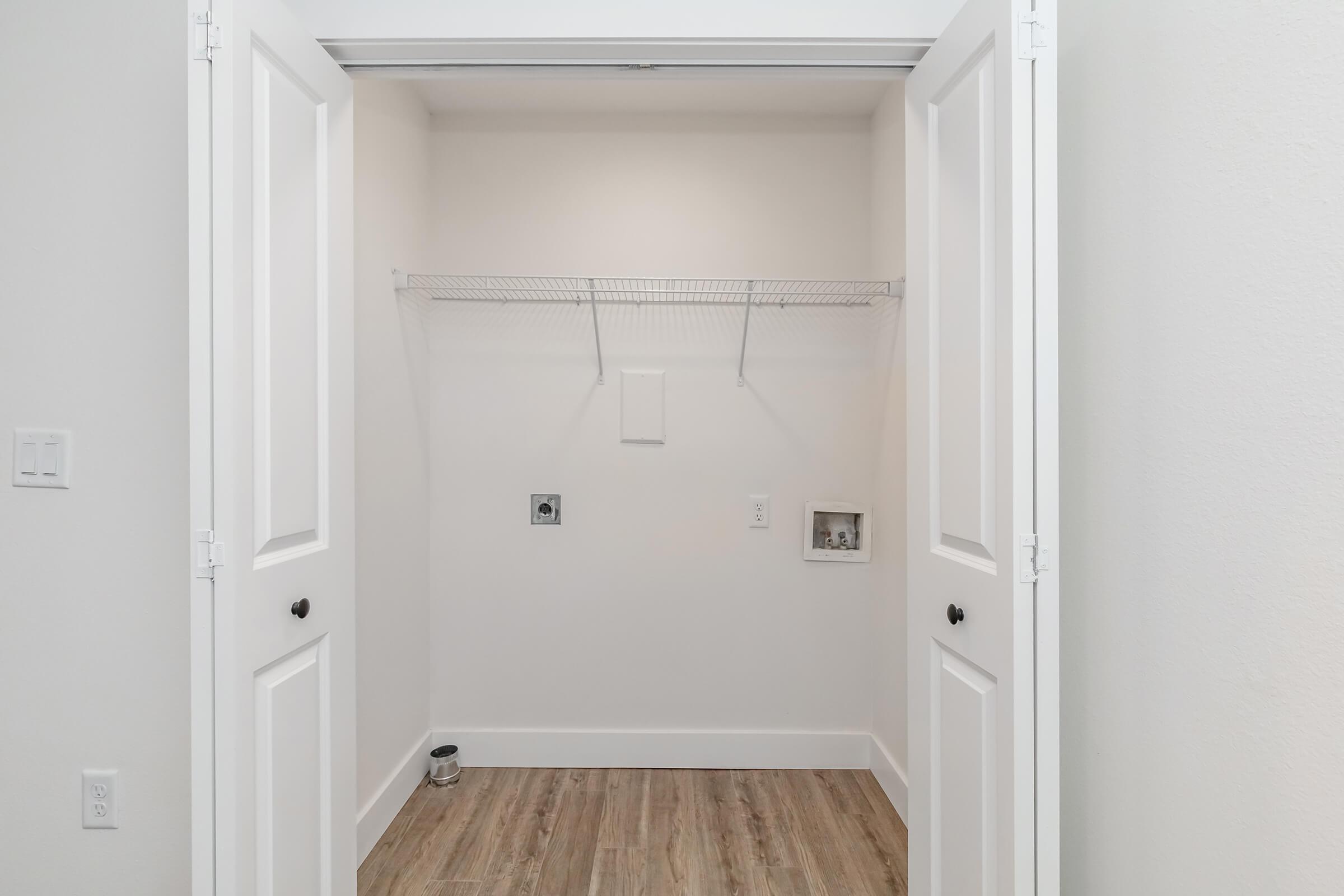 A spacious, empty closet featuring white double doors, light-colored walls, and wooden flooring. Shelving is installed on one wall, with electrical outlets visible. The closet is well-lit and uncluttered, providing a blank canvas for storage options.