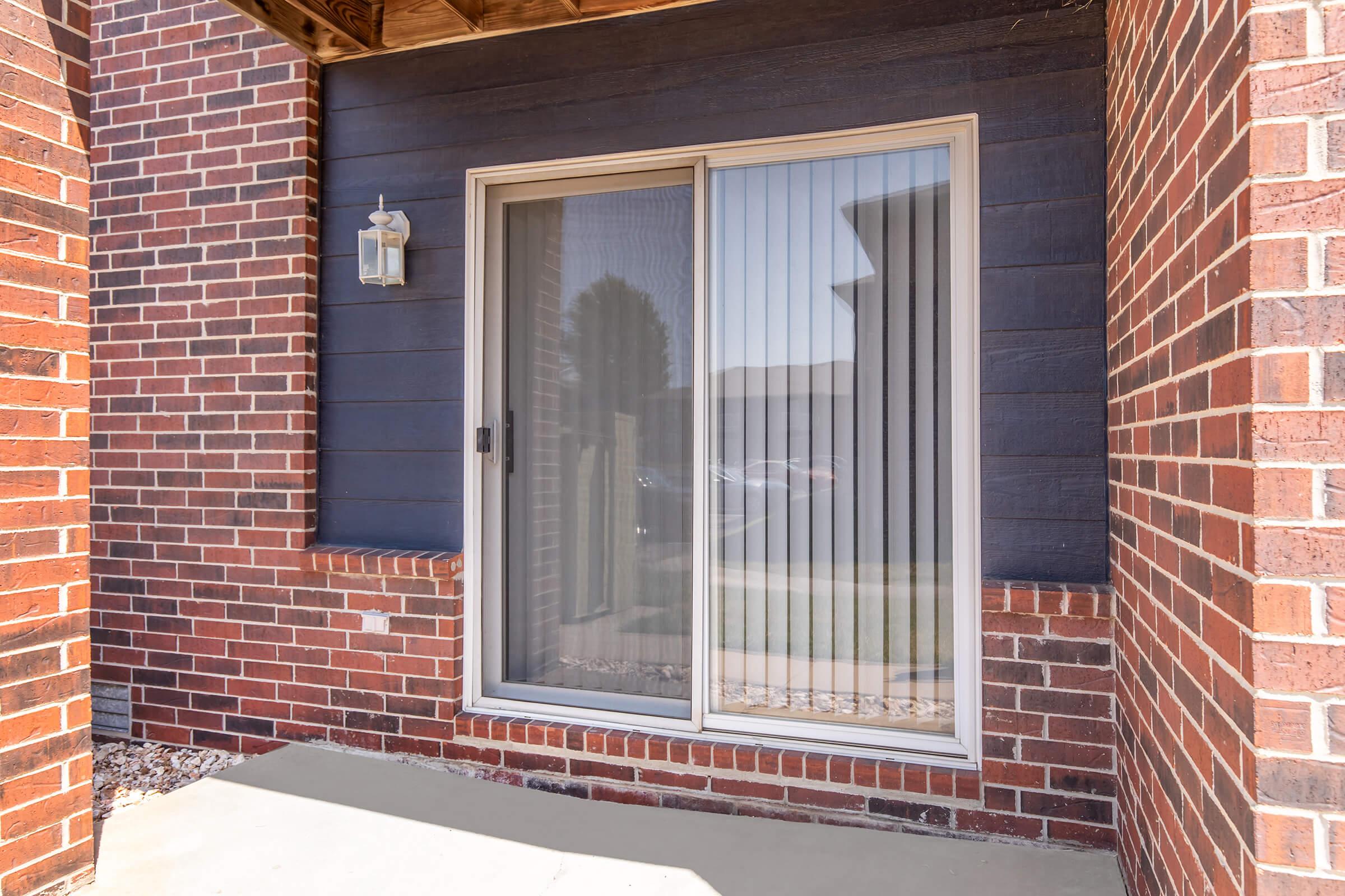 A sliding glass door with vertical blinds, set within a brick wall. The door is flanked by two brick columns and has a decorative light fixture above it. The surrounding area features a concrete base and landscaping visible in the background.