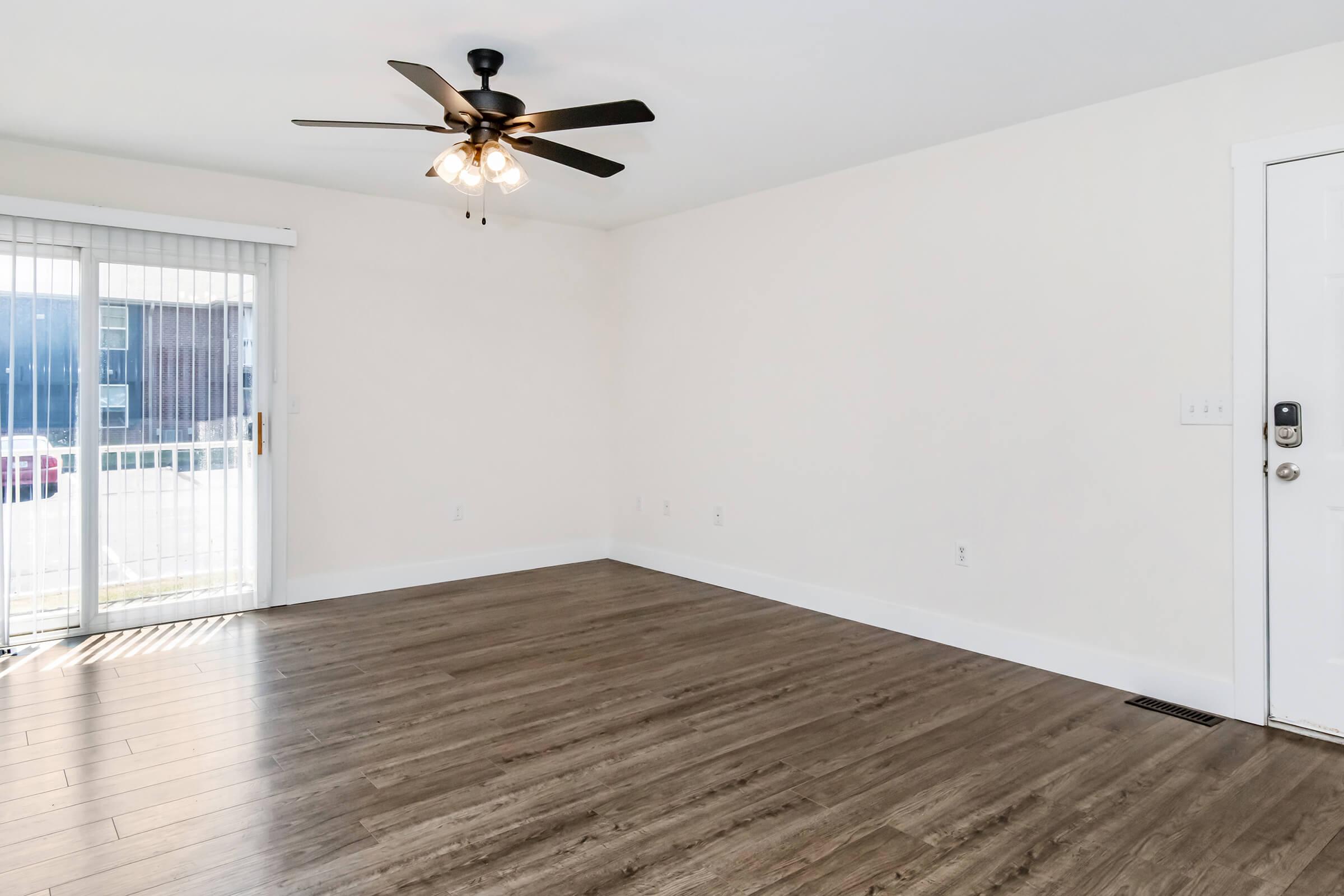 A spacious, empty room featuring light-colored walls, a dark wooden floor, and a ceiling fan with light fixtures. A sliding glass door leads to an outdoor area, allowing natural light to enter. The overall ambiance is bright and clean, suitable for new furnishings and decor.