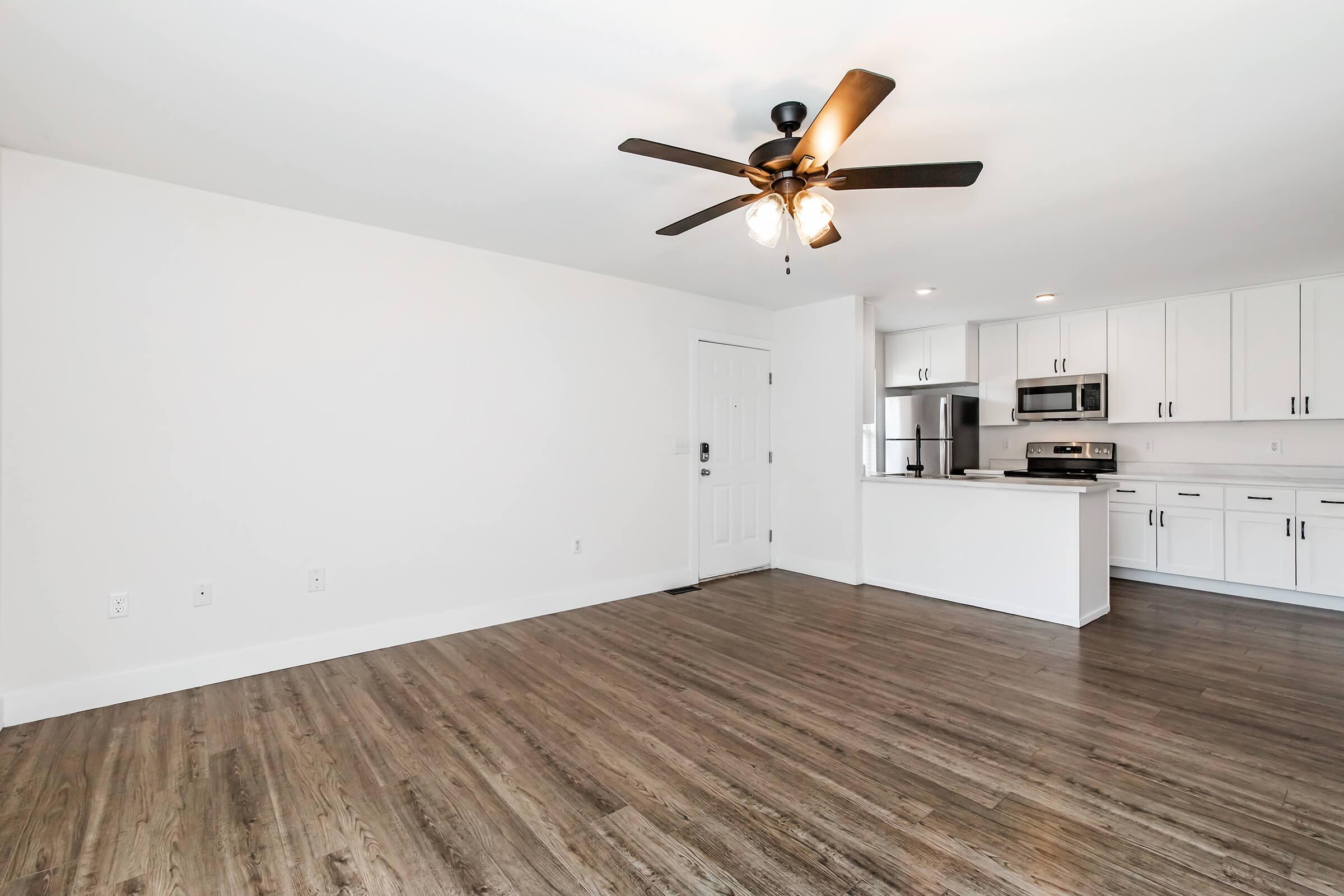 Spacious and modern living area with light hardwood flooring, white walls, and a ceiling fan. An open kitchen is visible, featuring white cabinetry and stainless steel appliances. A front door is located on the left side of the image, contributing to the bright, airy atmosphere of the room.