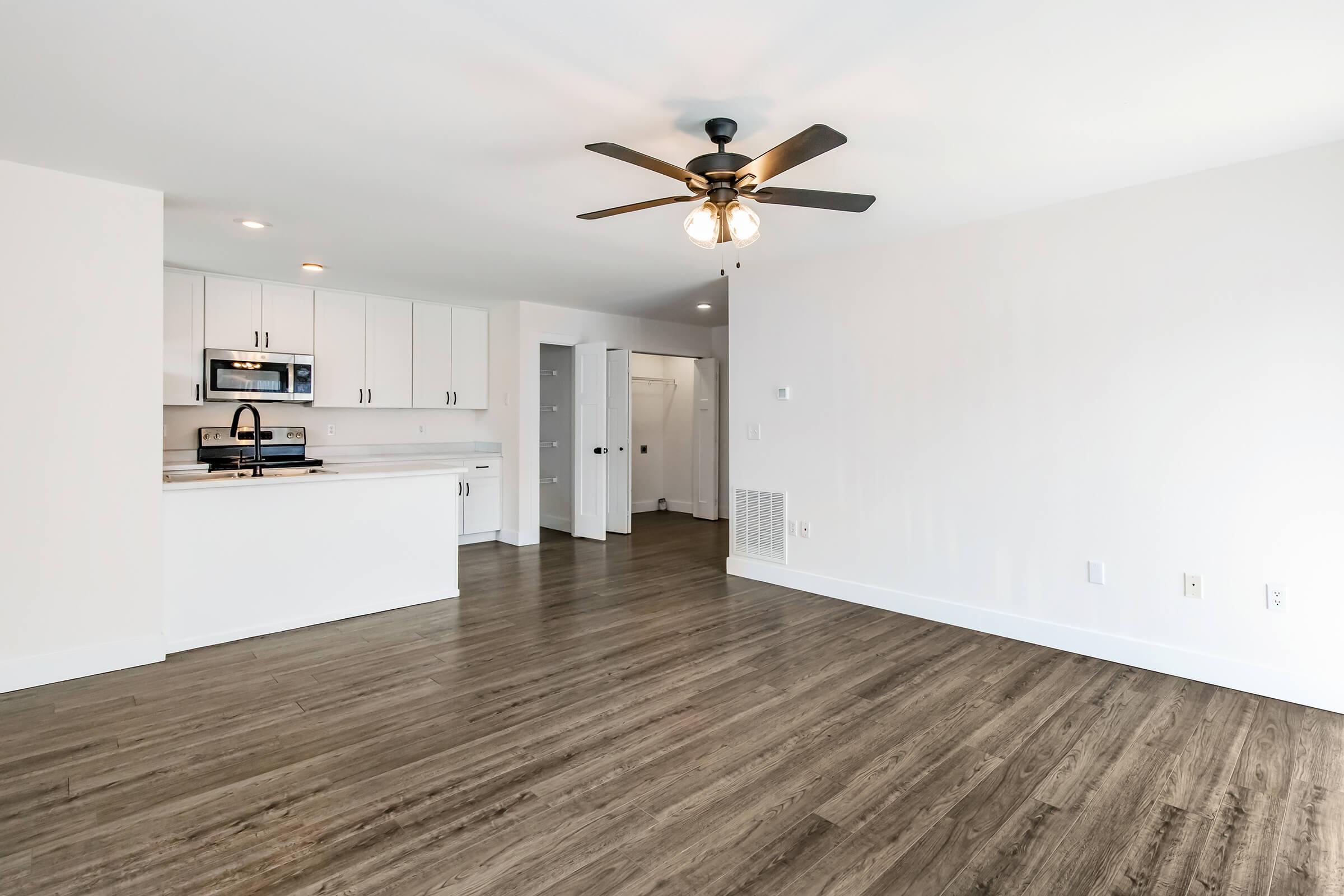 A spacious, modern living area featuring a ceiling fan, hardwood-style flooring, and a view of an open kitchen with white cabinetry and stainless steel appliances. The walls are painted light colors, creating a bright and inviting atmosphere. A hallway leads to other rooms.