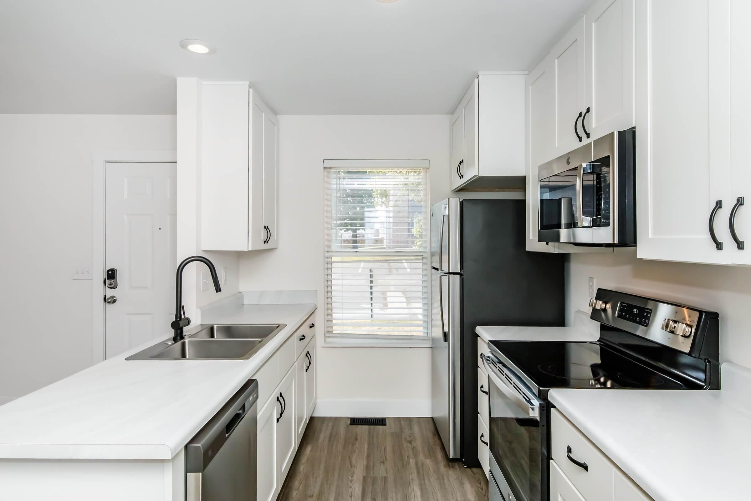 A modern kitchen featuring white cabinets, stainless steel appliances, and a double sink. The space includes a black stove, microwave, and refrigerator, with natural light coming through a window. The flooring is light wood, and the walls are painted white, creating a bright and clean aesthetic.