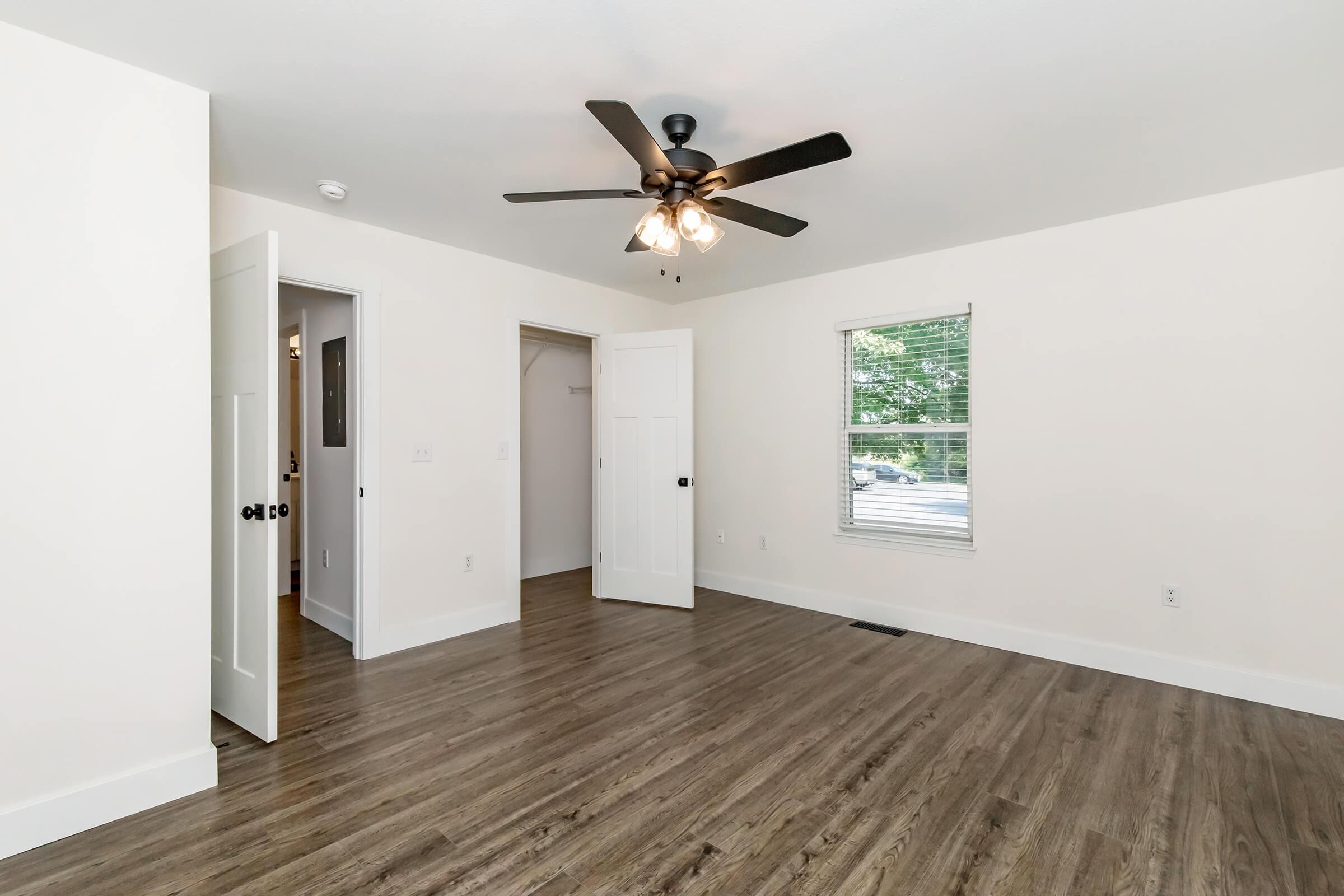 A bright, empty room with light-colored walls and a ceiling fan. The room features a wood-like floor, a window with natural light, and two open doors leading to other spaces. The overall atmosphere is clean and modern, ideal for decorating or furnishing.