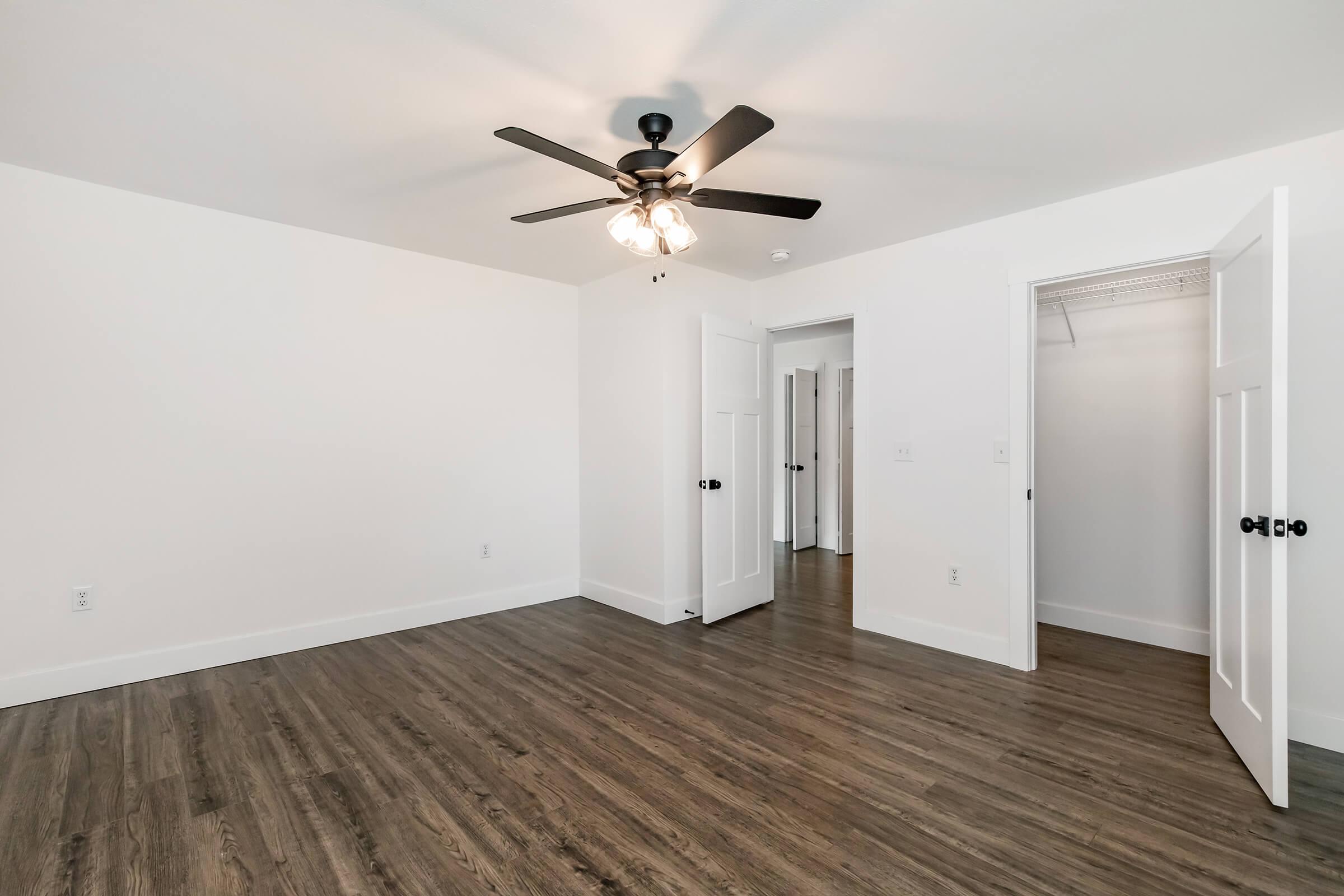 A spacious, vacant room with a ceiling fan, featuring light-colored walls and laminate flooring. Two white doors are open, revealing a closet and a doorway leading to another room. The floor is clean and polished, creating a bright and airy atmosphere.