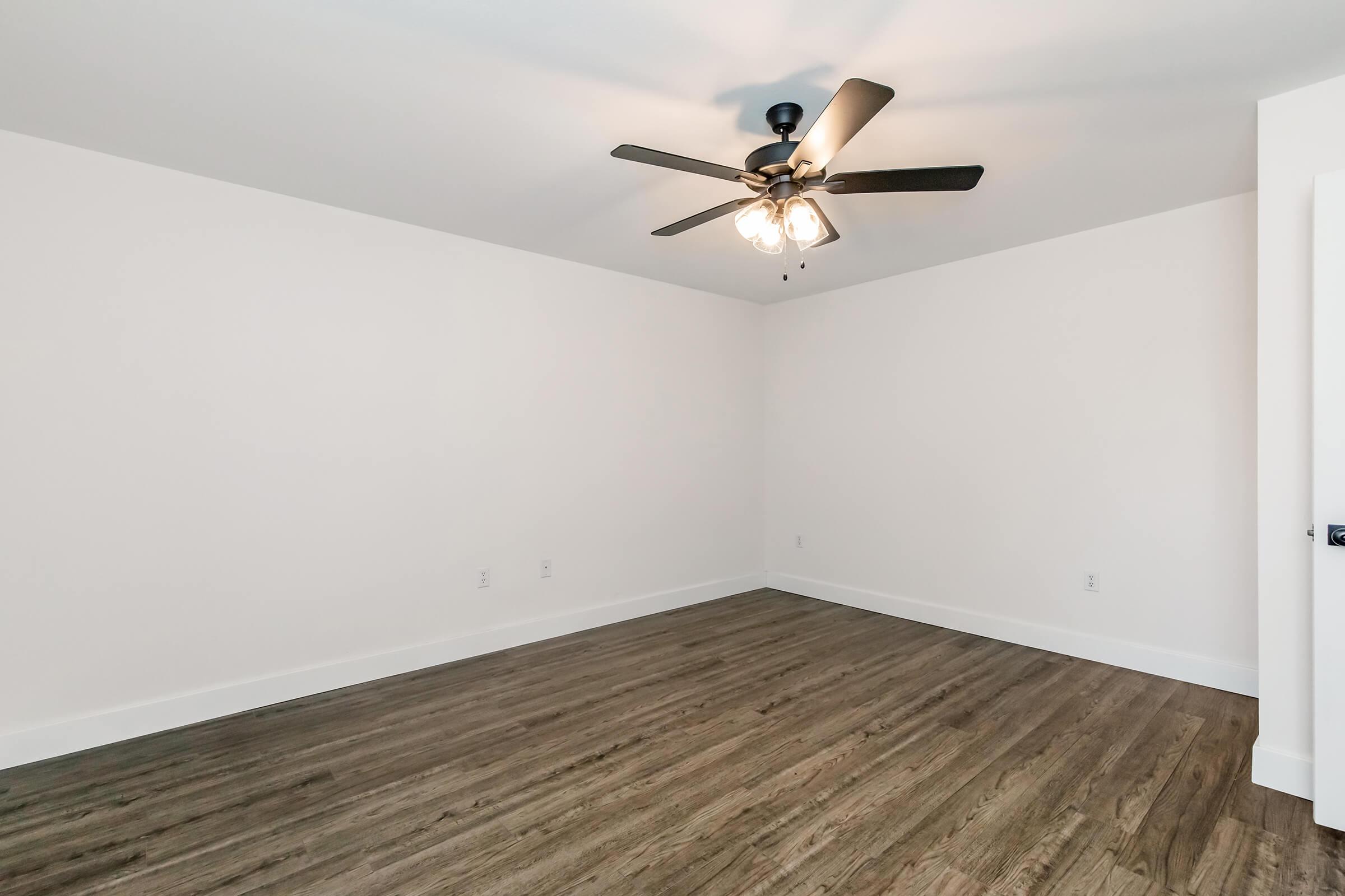 Empty room with light wood flooring and white walls. A ceiling fan with three light fixtures is centered in the room, and there are no furnishings. The space appears bright and spacious, creating an inviting atmosphere.