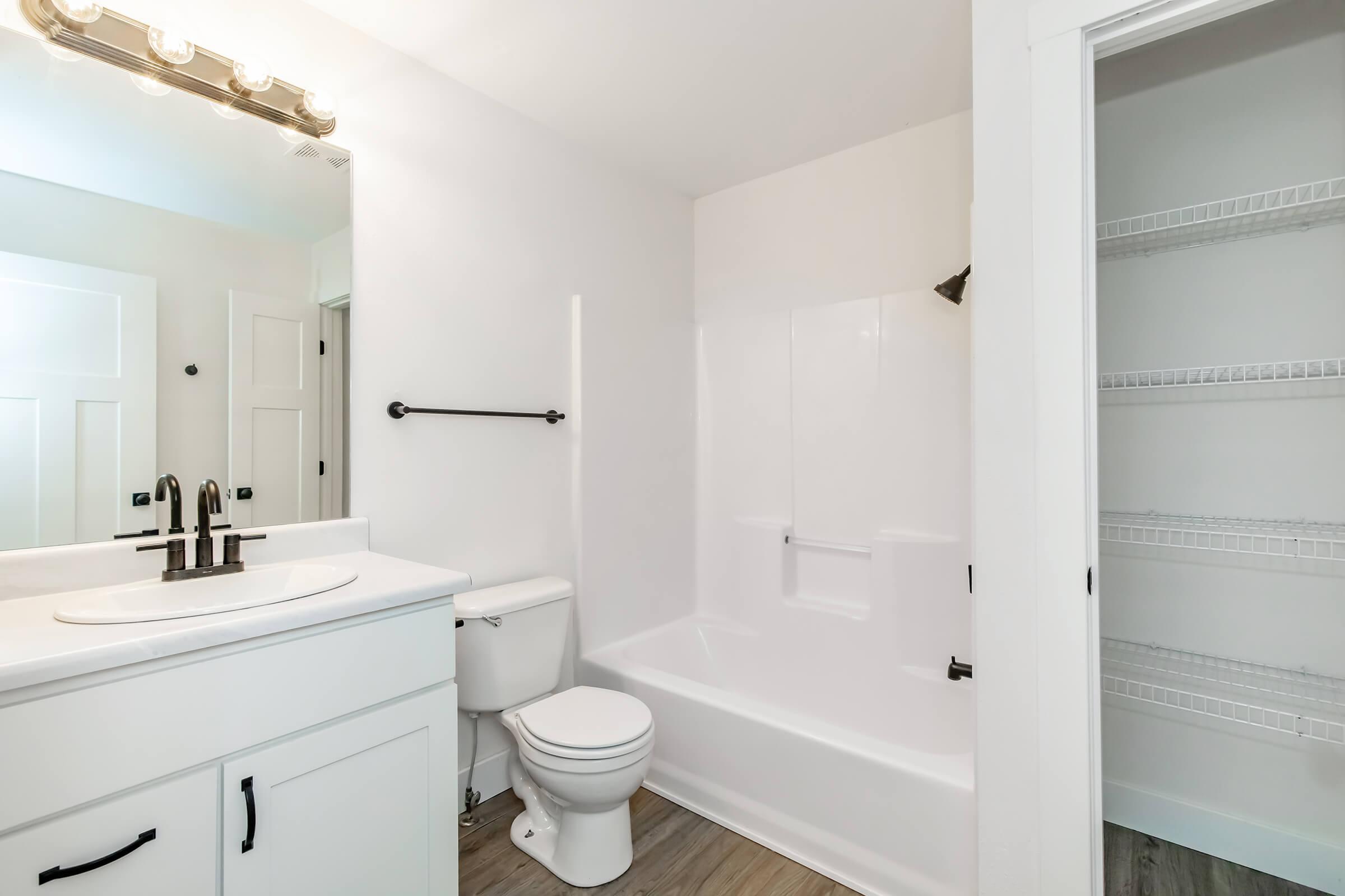 A clean, modern bathroom featuring a white bathtub and shower combination, a white vanity with a round sink and faucet, a toilet, and a mirrored light fixture above the sink. To the right, there is a closet with shelves. The walls are painted white, and the flooring is a light wood style.