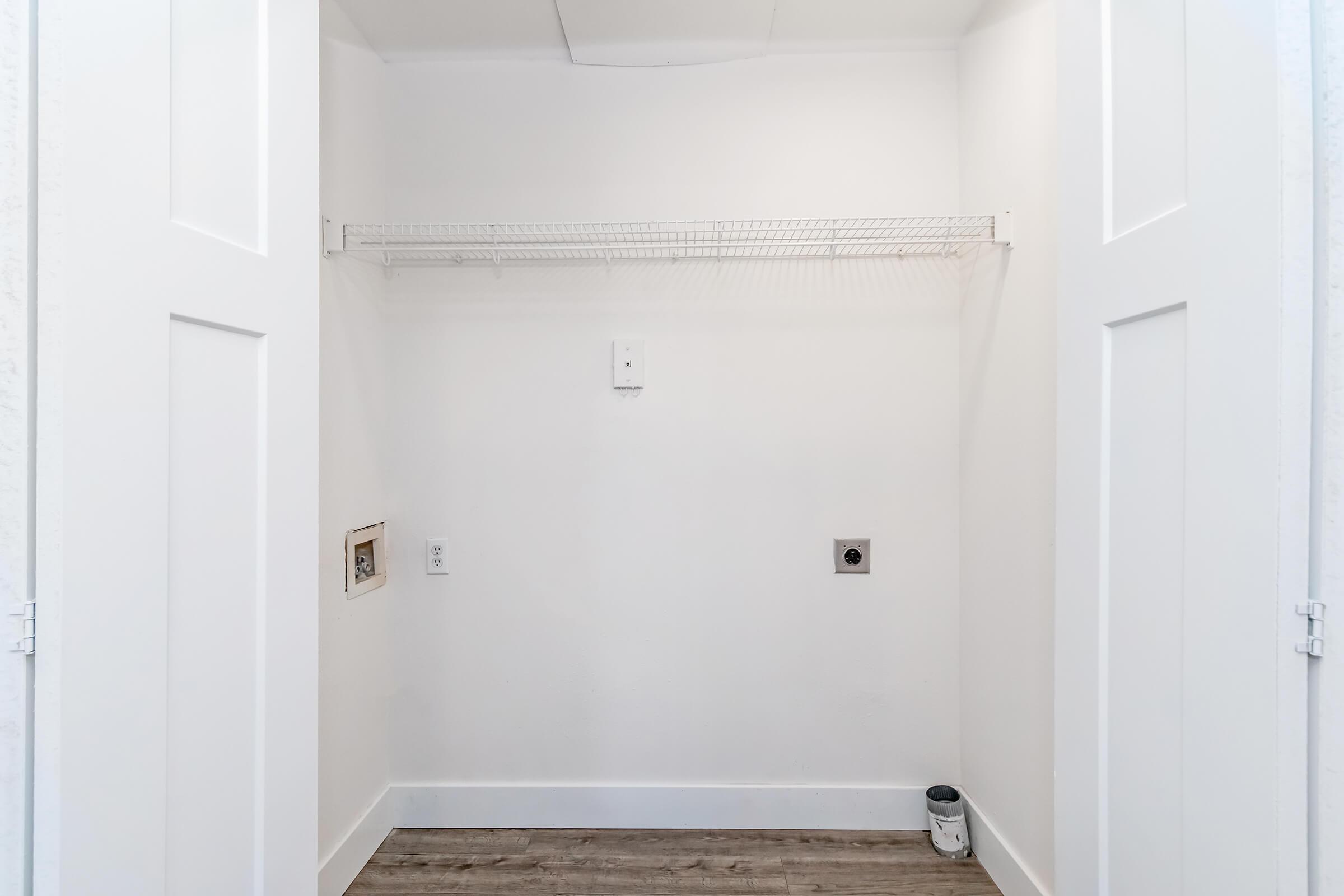 Empty closet space with white walls, a wire shelf mounted on the back wall, and a small appliance outlet. The floor is covered with light-colored laminate, and there is a small container on the floor. The closet doors are open, revealing the minimalistic interior.